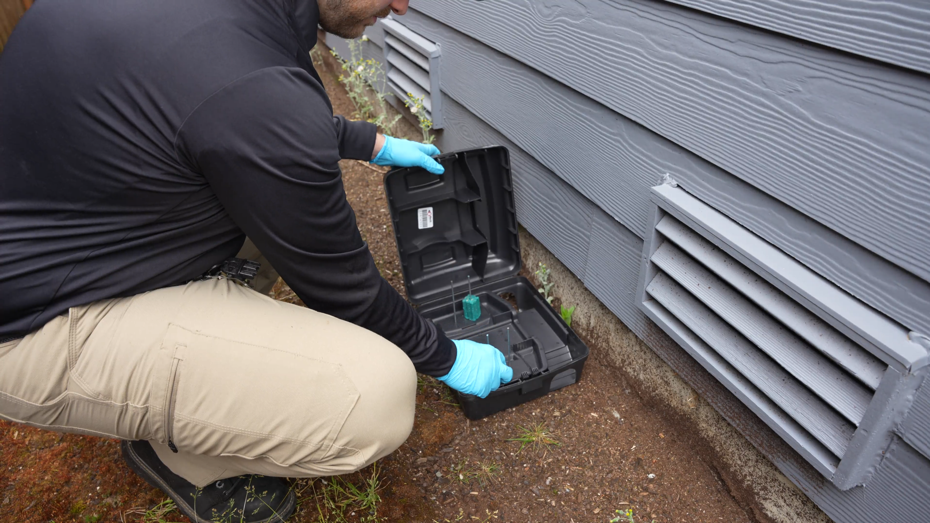 Worker in blue gloves inspecting an open black trap beside a gray house vent.