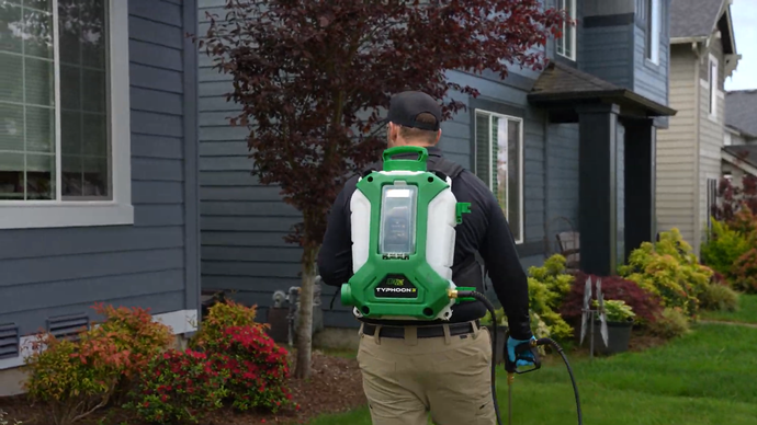 Person wearing a bright green backpack leaf blower works along a house-lined walkway.