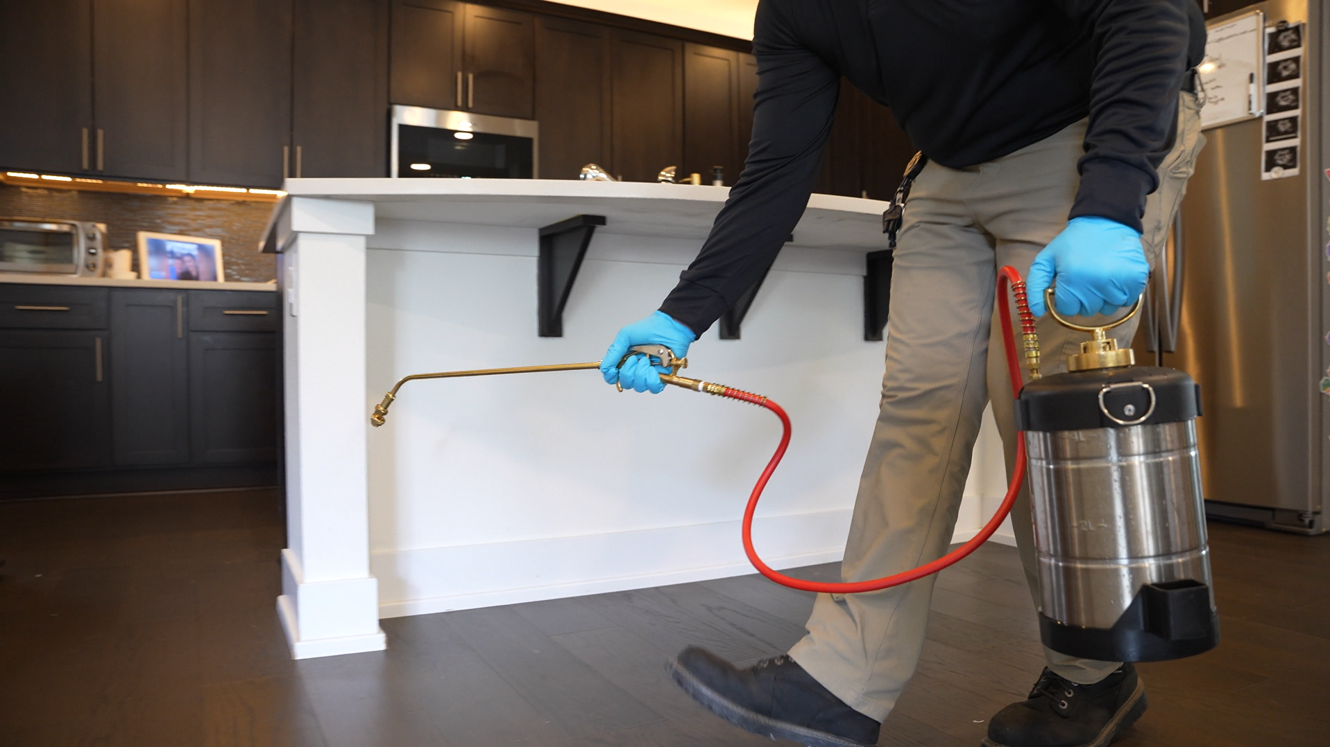 Person in blue gloves uses a red hose to clean under a kitchen counter with a metal canister vacuum.
