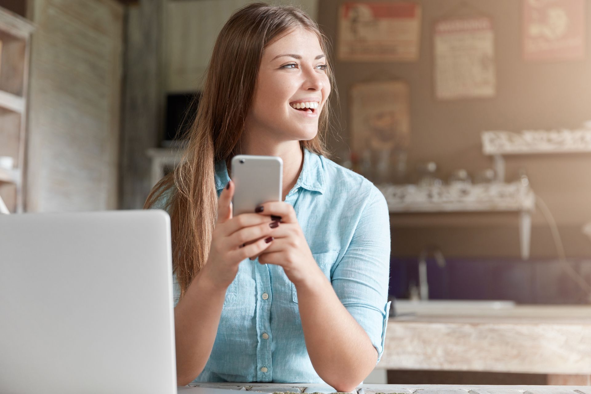Woman smiling and holding a smartphone beside a laptop in a cafe