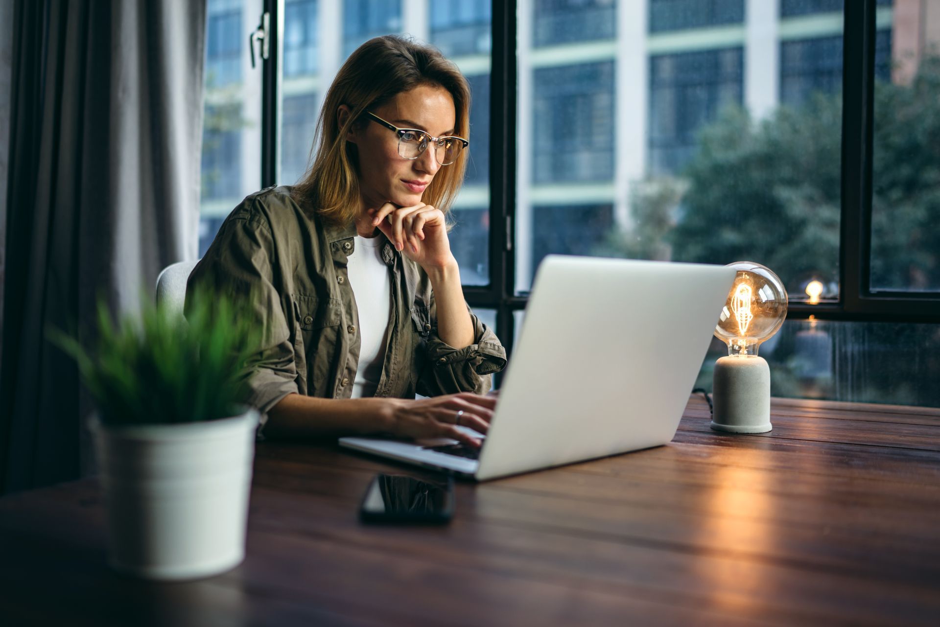 Person working on a laptop at a wooden desk by a window, with a potted plant and lit lamp nearby.