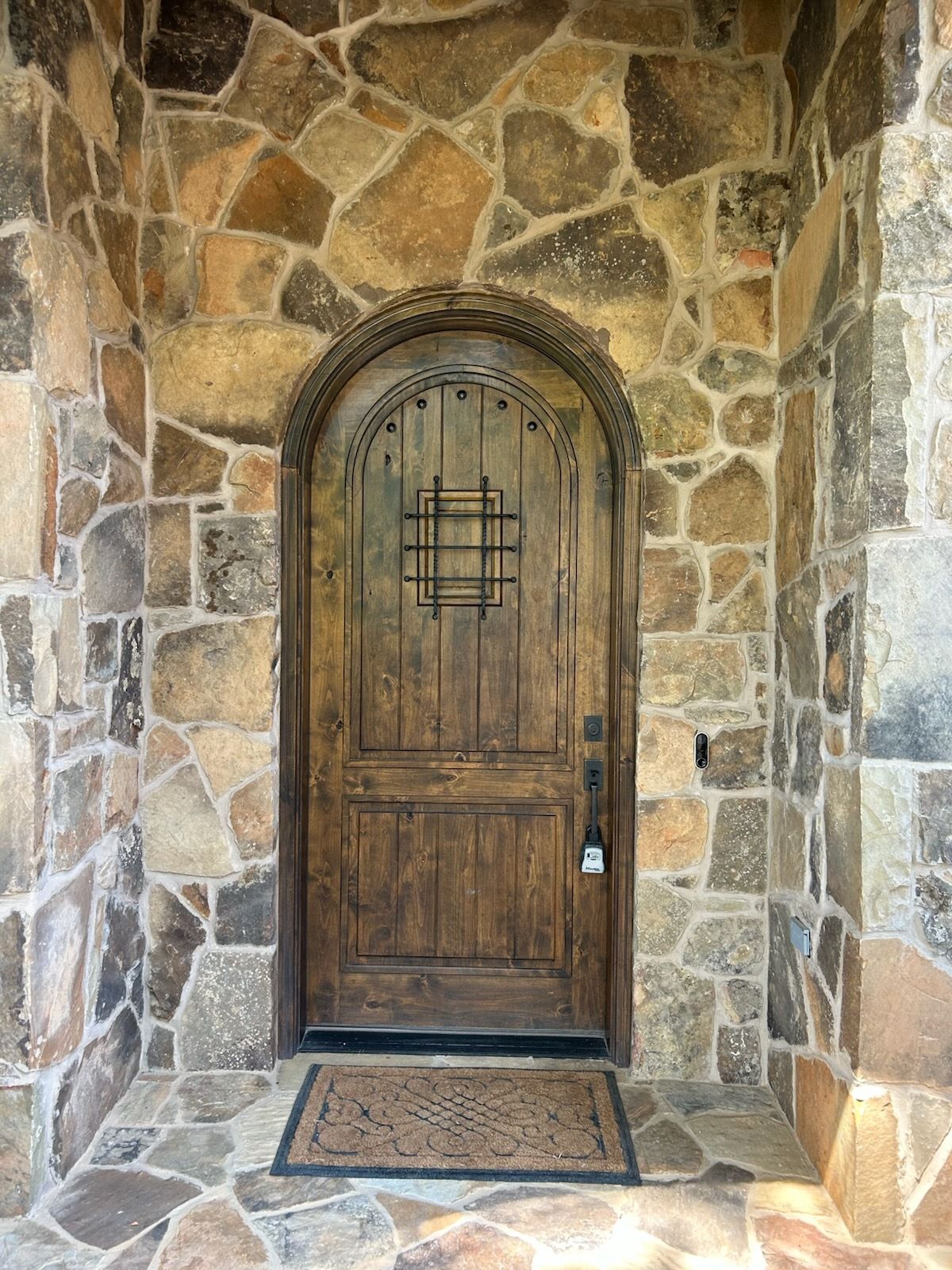 Wooden arched front door set in a stone wall with a doormat and small side light.