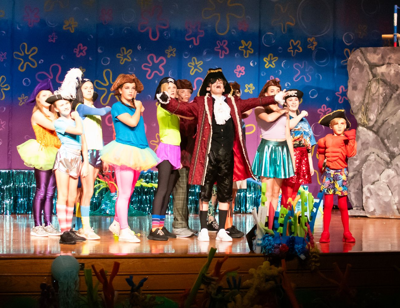 A stage production with costumed characters. A pirate commands a group of colorful sea creatures.