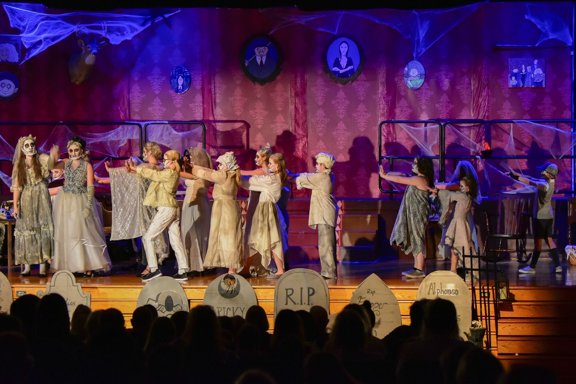 Stage performance with costumed actors, graveyard backdrop, and audience in a theater.
