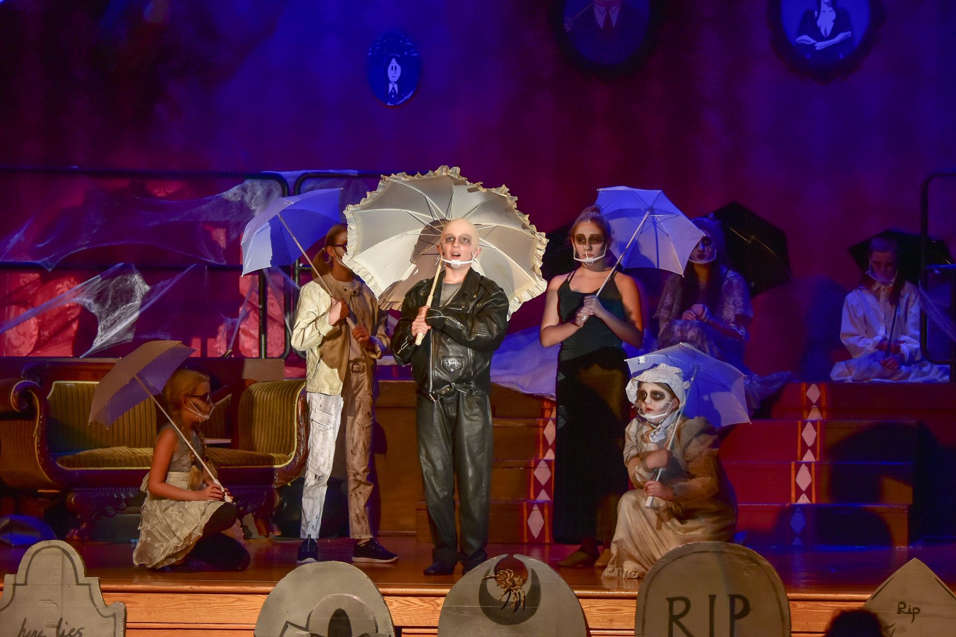 Actors in a spooky stage production, dressed as ghosts and ghouls, holding umbrellas and posing on a stage.