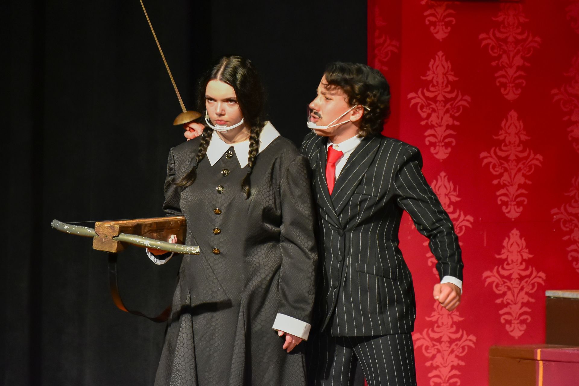 Actors in Addams Family costumes: Wednesday with crossbow, boy in pinstripes, red backdrop.
