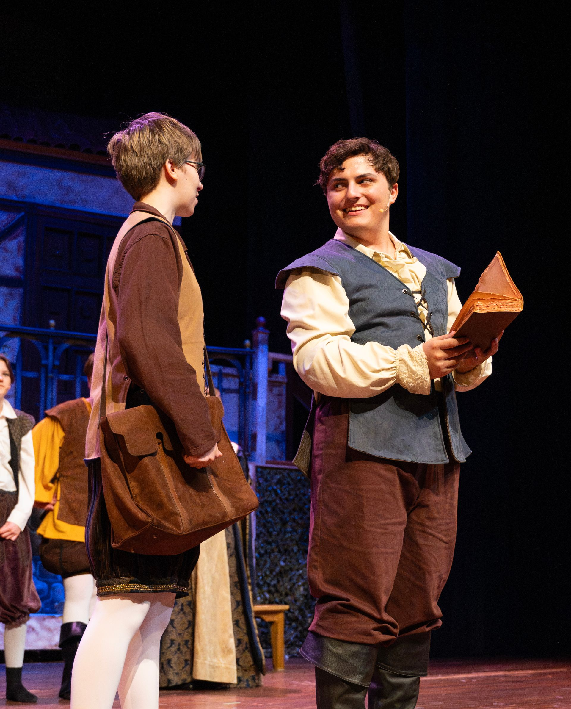 Two actors in Renaissance costumes on stage, one holding a book, smiling.