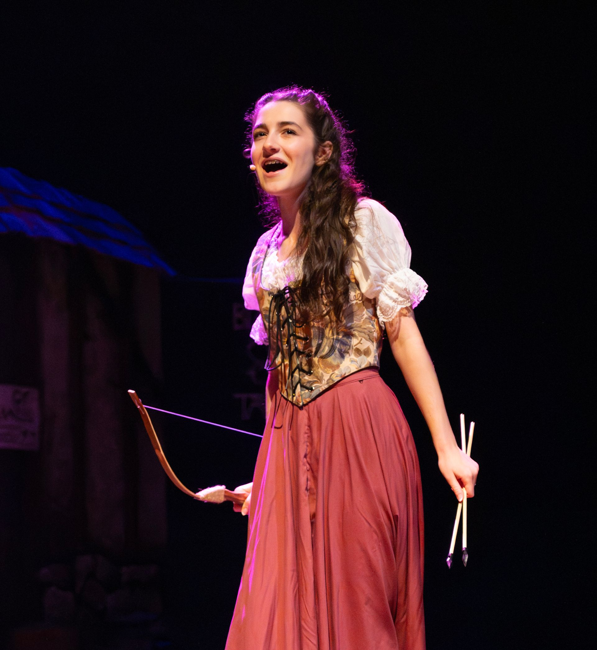 Woman in costume on stage, holding bow and arrows, singing, red skirt, white blouse.