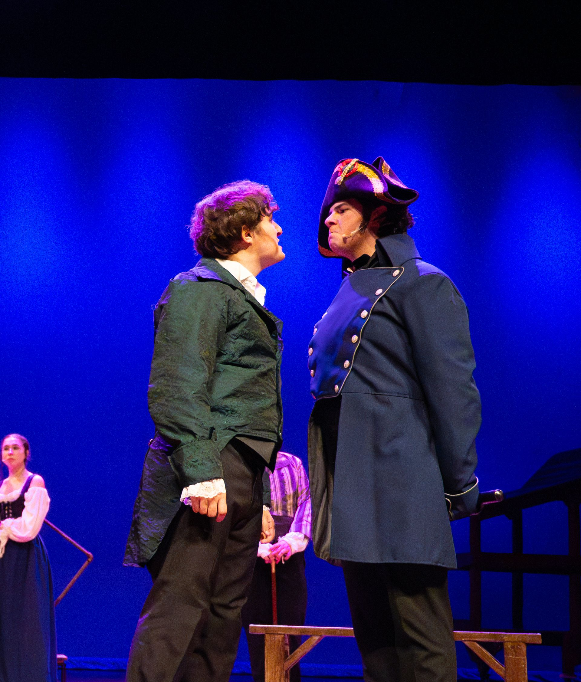 Two actors face each other onstage; one in green jacket, other in blue coat. Blue backdrop.