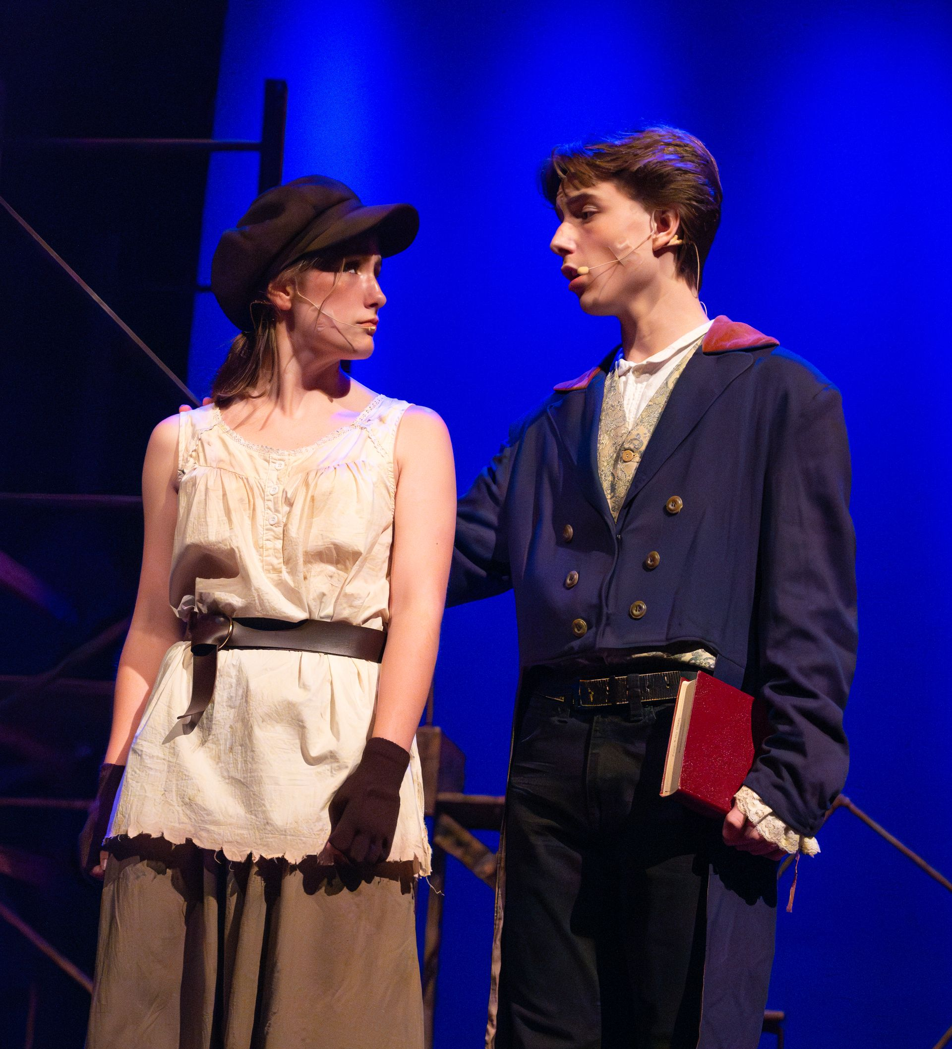 Two people in period costumes on stage. Woman with a cap looks on, man with a book gestures with his right hand.