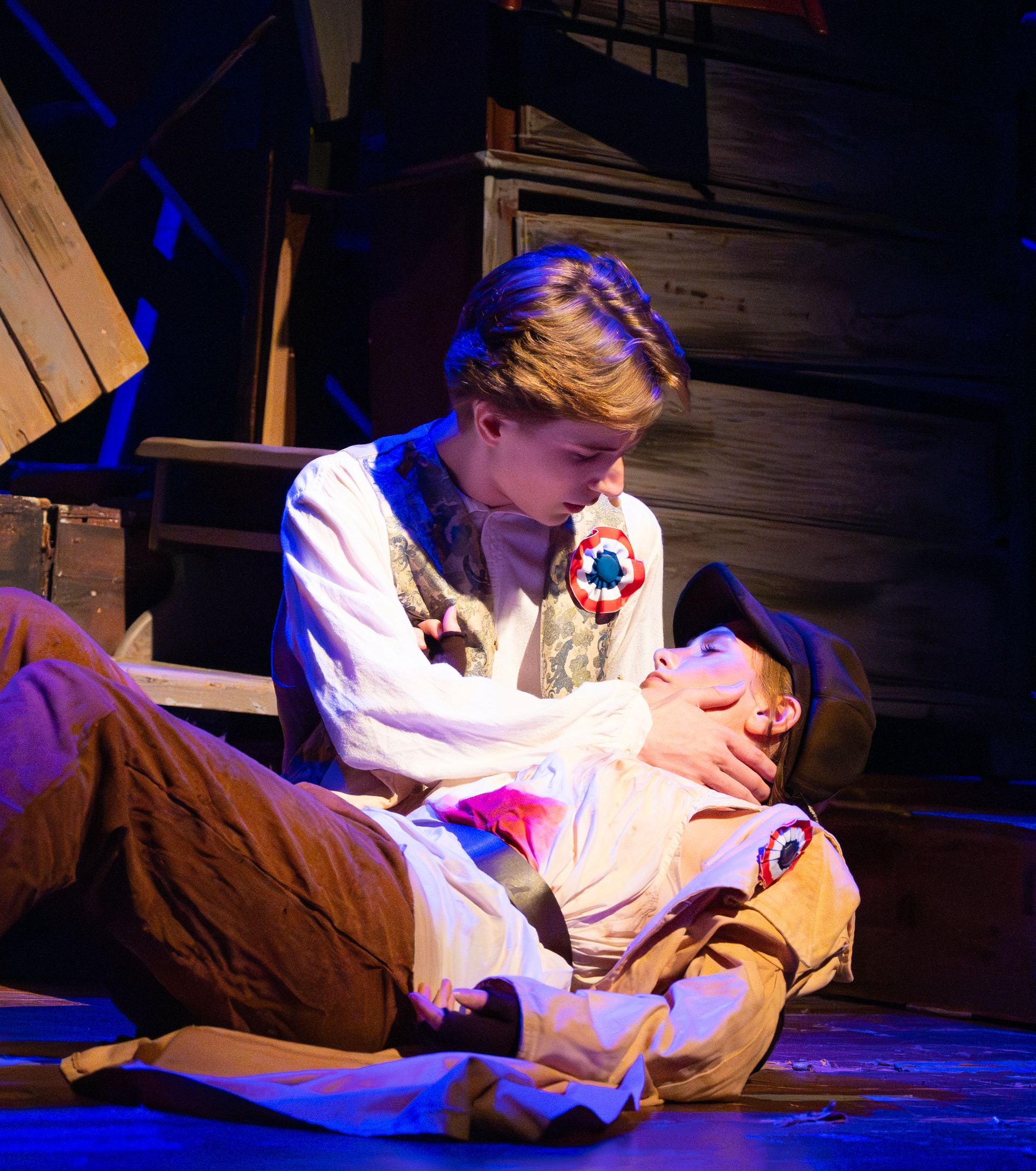 Two young men, one lying injured, is held by the other, concerned. A stage set with wood, blue light.