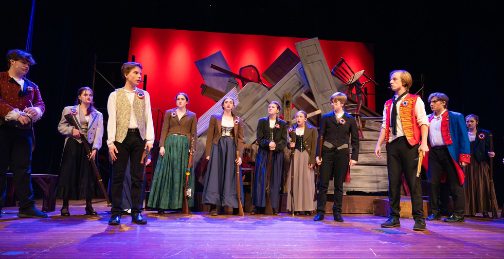 A stage production: actors in period costumes, posed on a stage with a red backdrop.