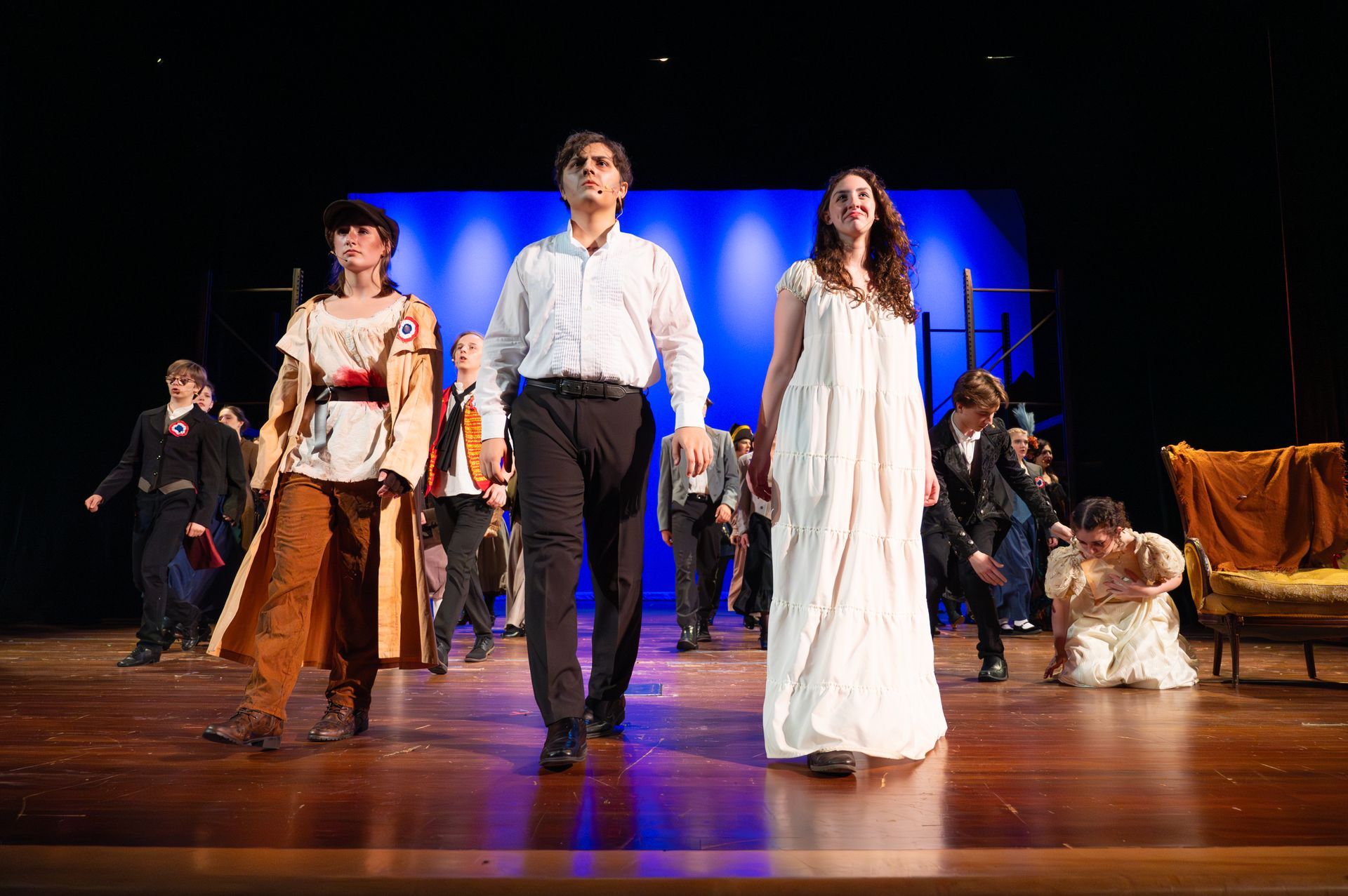 A play performance: Three actors in costume walk forward, flanked by others on a stage with a blue backdrop.