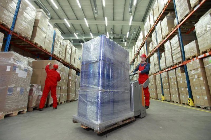 A man is pushing a cart with a large item wrapped in plastic in a warehouse.