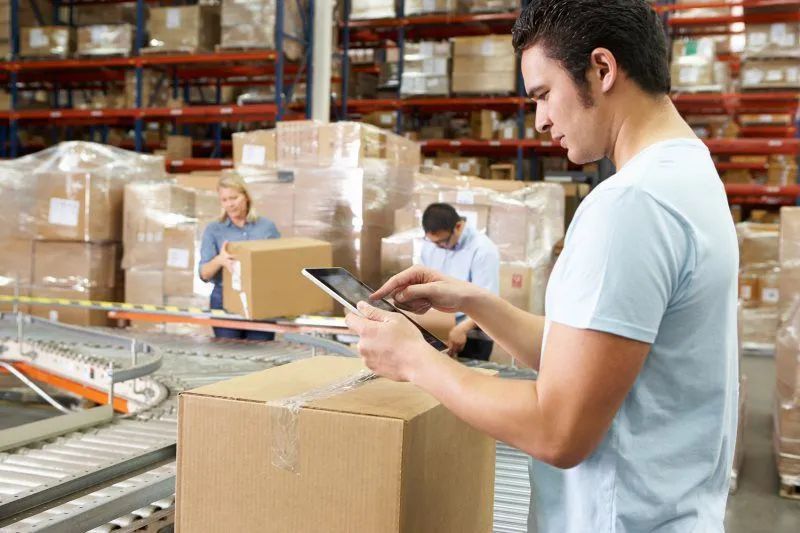 A man is using a tablet in a warehouse while looking at a box.