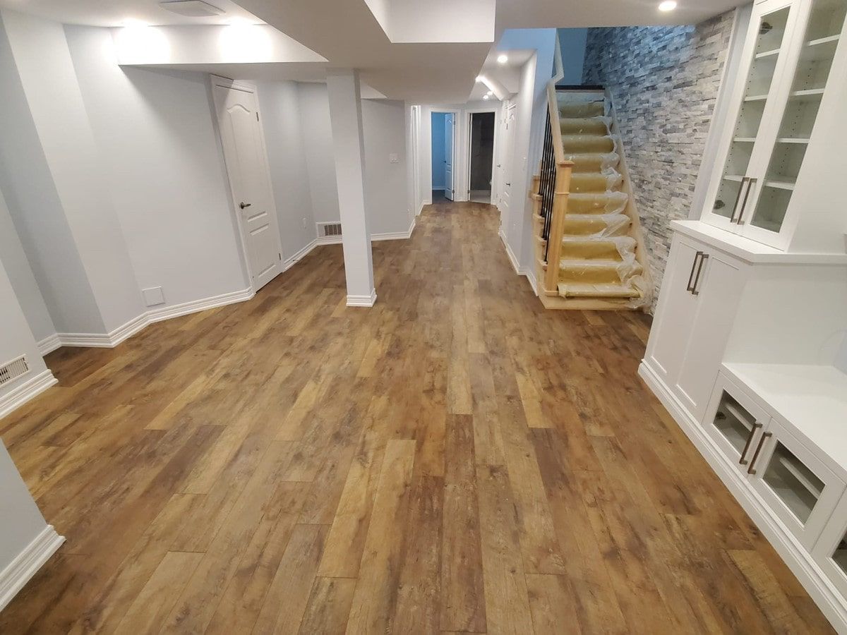 Spacious basement interior with wood-look flooring, white walls, and stairs with beige carpet leading up.