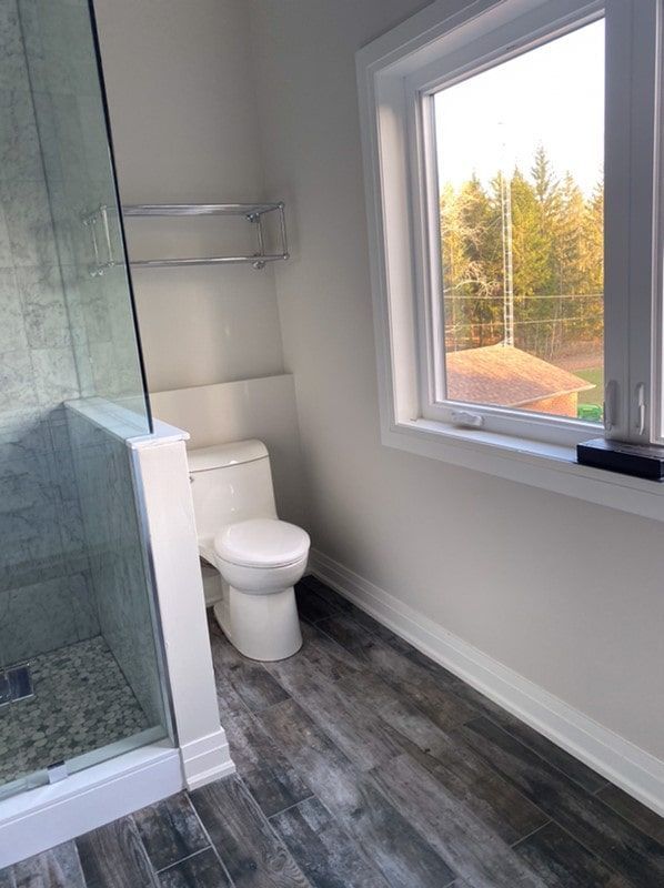 Bathroom with shower, toilet, and window, featuring dark wood-look flooring and light-colored walls.