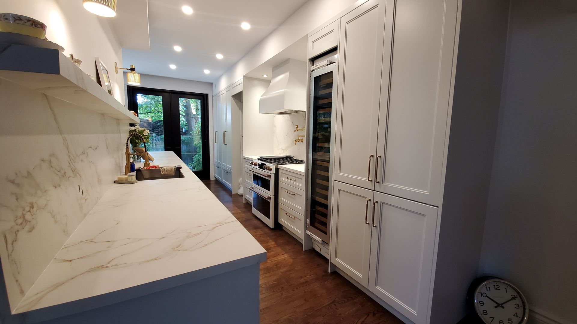 Long, narrow, white kitchen with marble countertop, dark wood floors, and black door to outside.