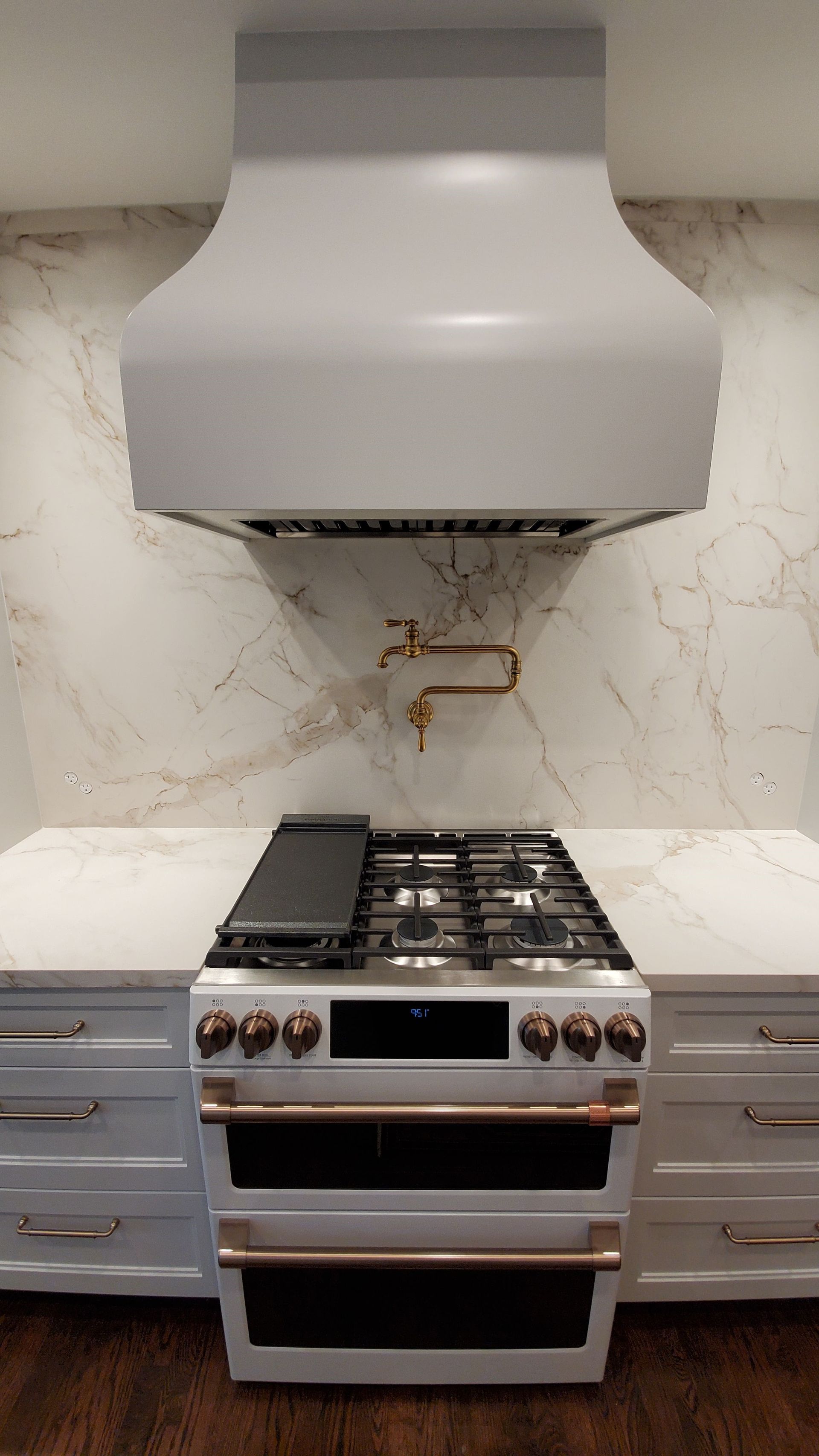 White kitchen stove with bronze accents under a grey range hood and marble backsplash.