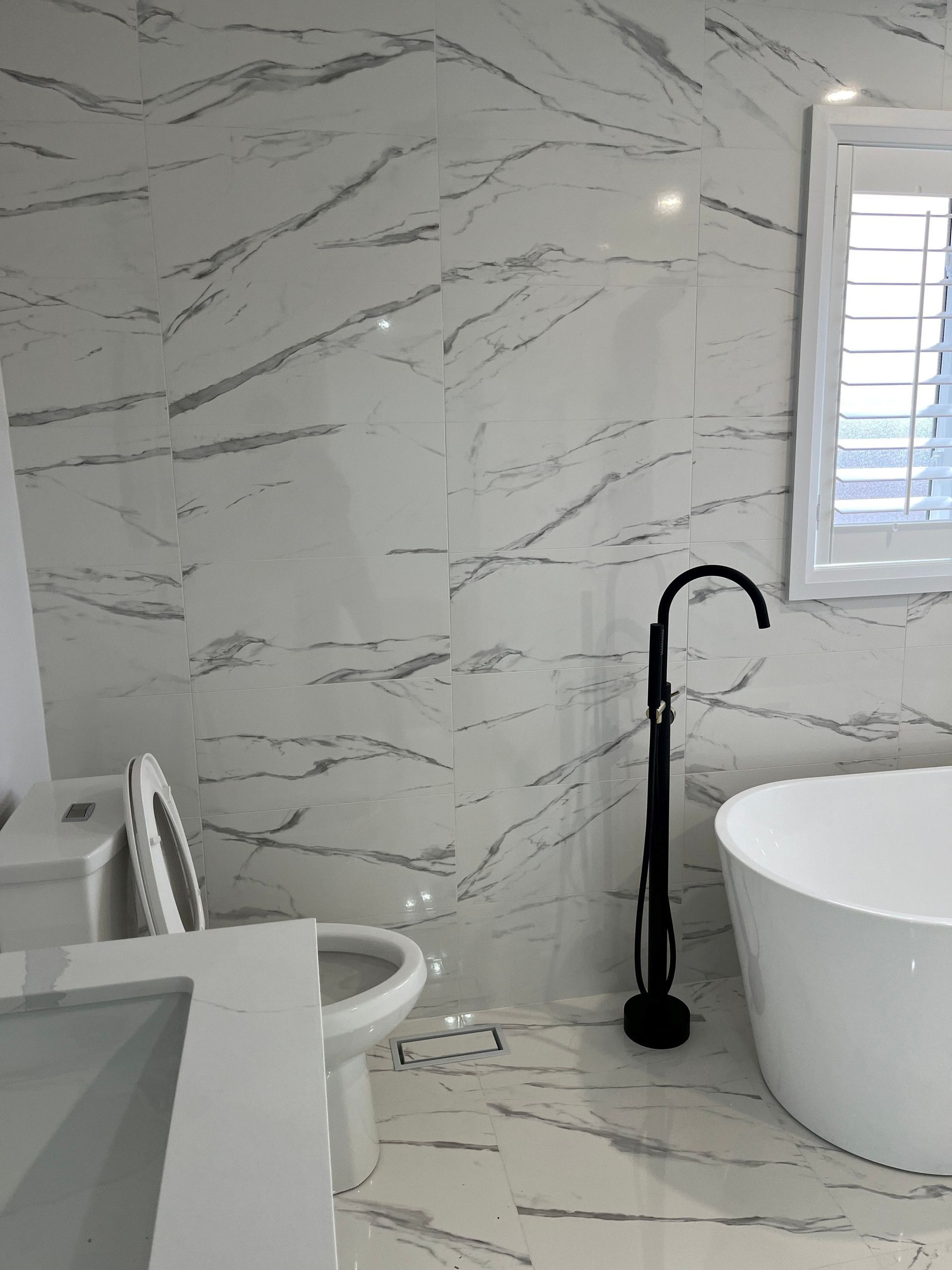 Bathroom with white marble-look tile, toilet, sink, bathtub, and black faucet. A window with shutters is on the right.
