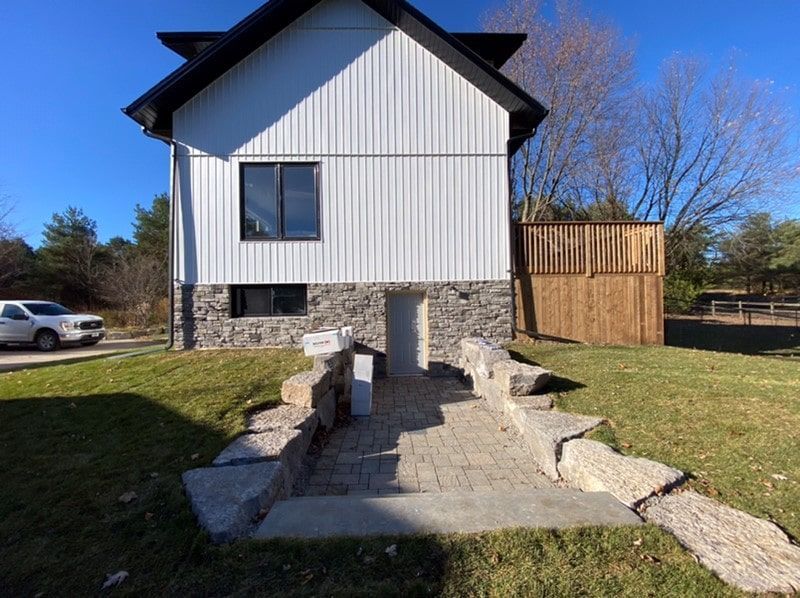 Side view of a house with stone base, white siding, and a pathway leading to a doorway.