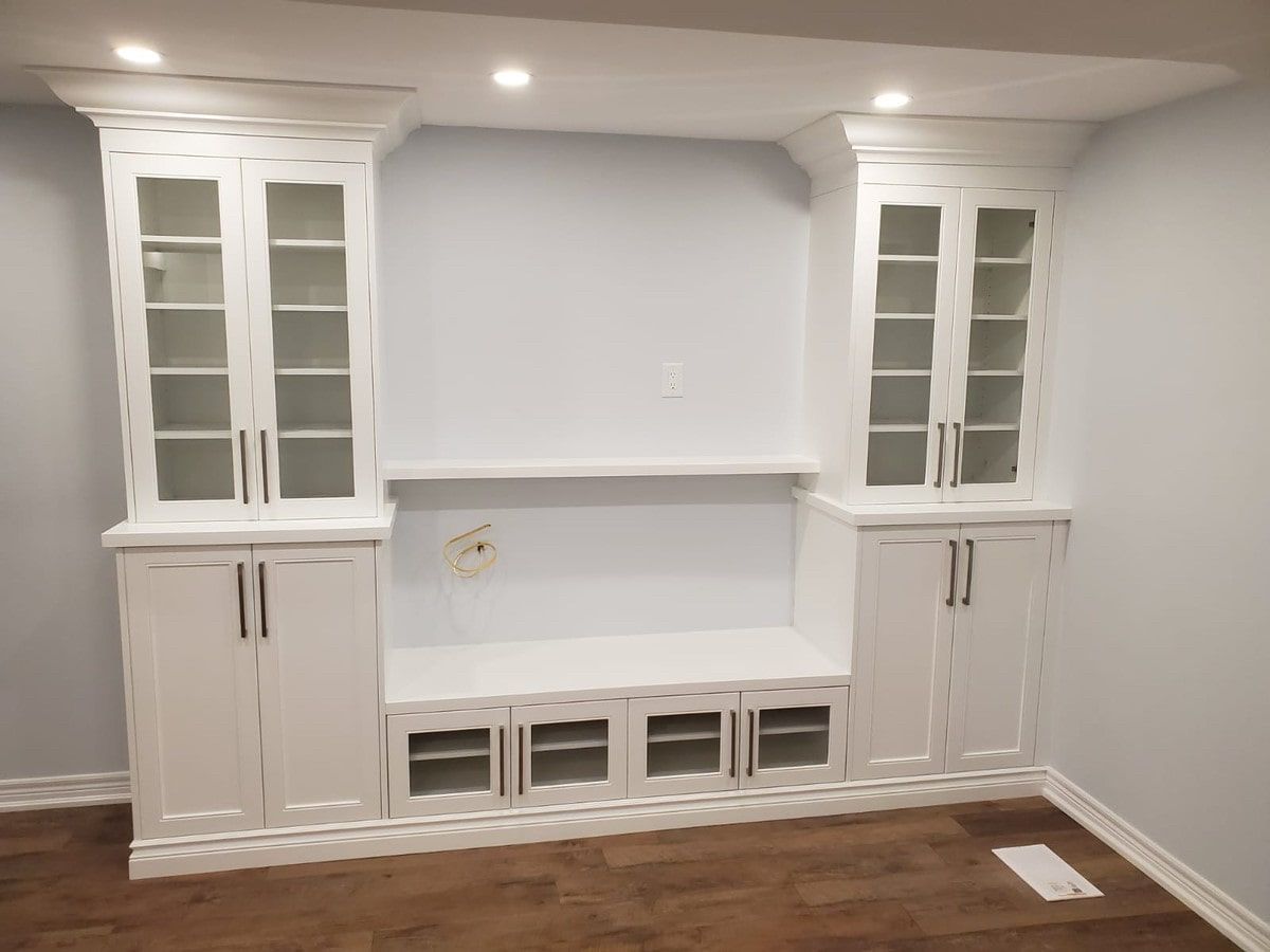 White built-in cabinet unit with glass-fronted upper cabinets, lower storage, and a central shelf against a light blue wall.