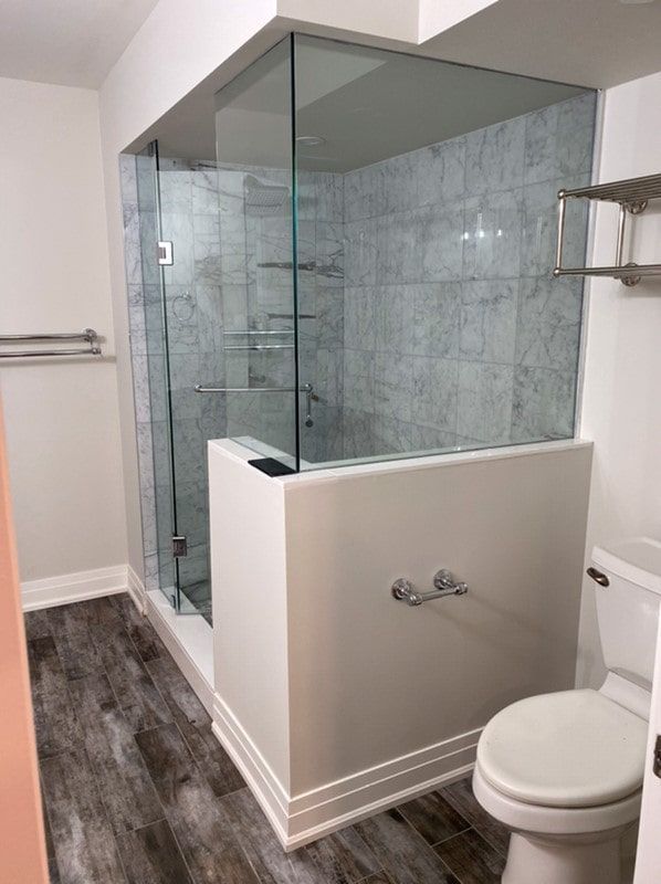 Modern bathroom with glass-enclosed shower, gray tile, white walls and trim, wood-look flooring, and a toilet.