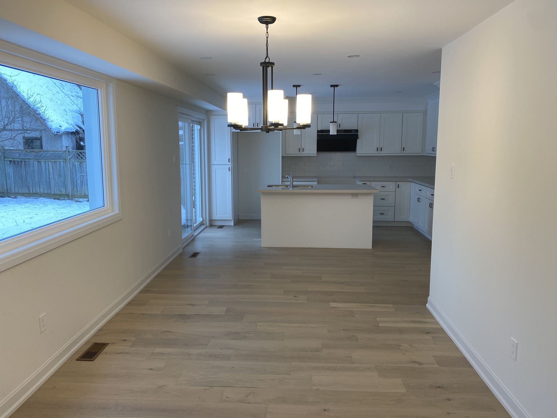 Dining area with a kitchen in the background. Light wood floors, white walls, and a pendant light fixture.