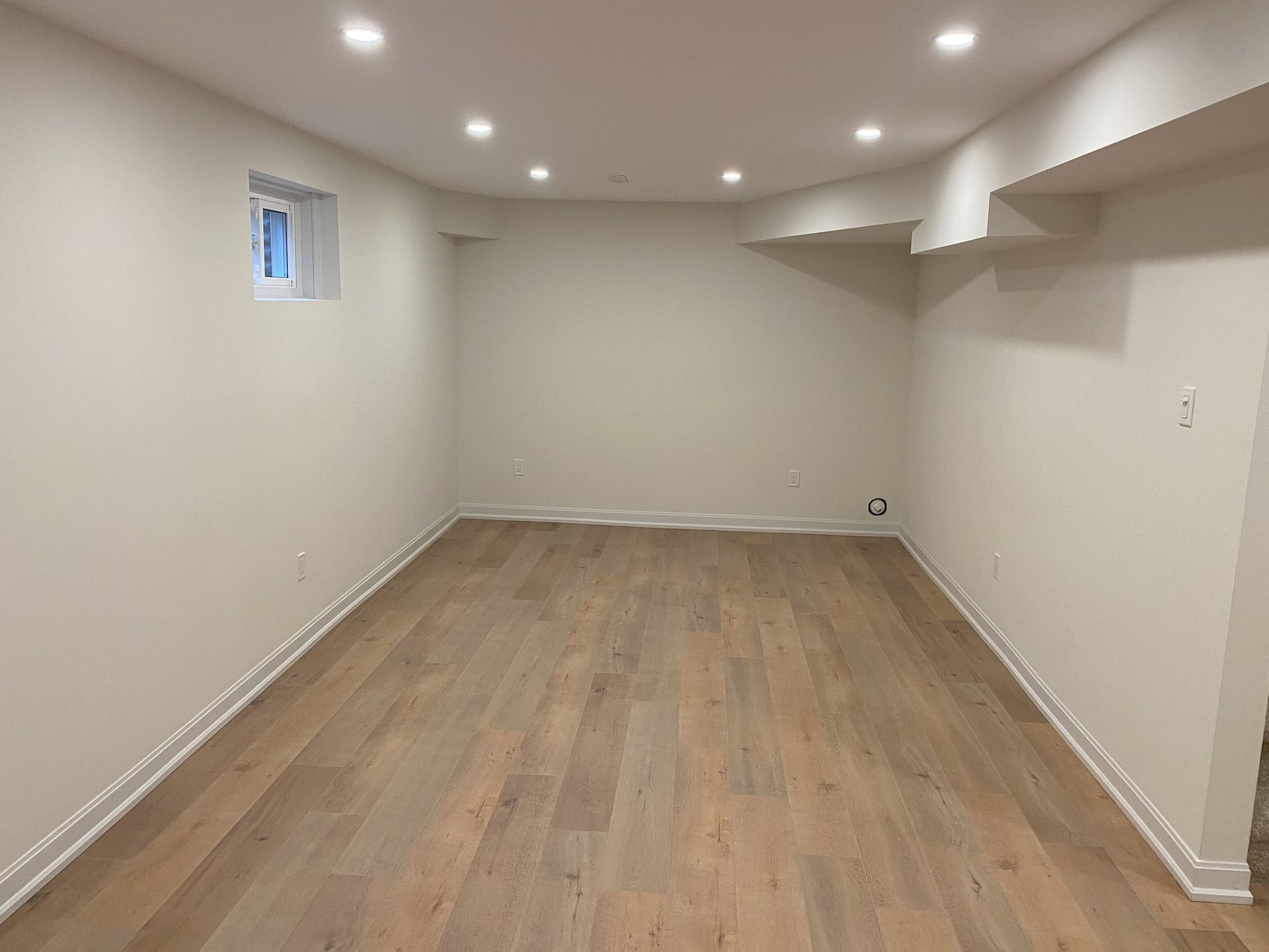 Empty room with light wood flooring, white walls, recessed ceiling lights, and small window.