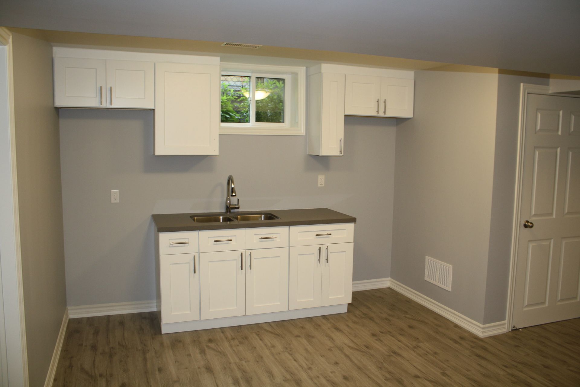 White kitchen cabinets and sink in a basement, gray walls, small window, and wood-look flooring.