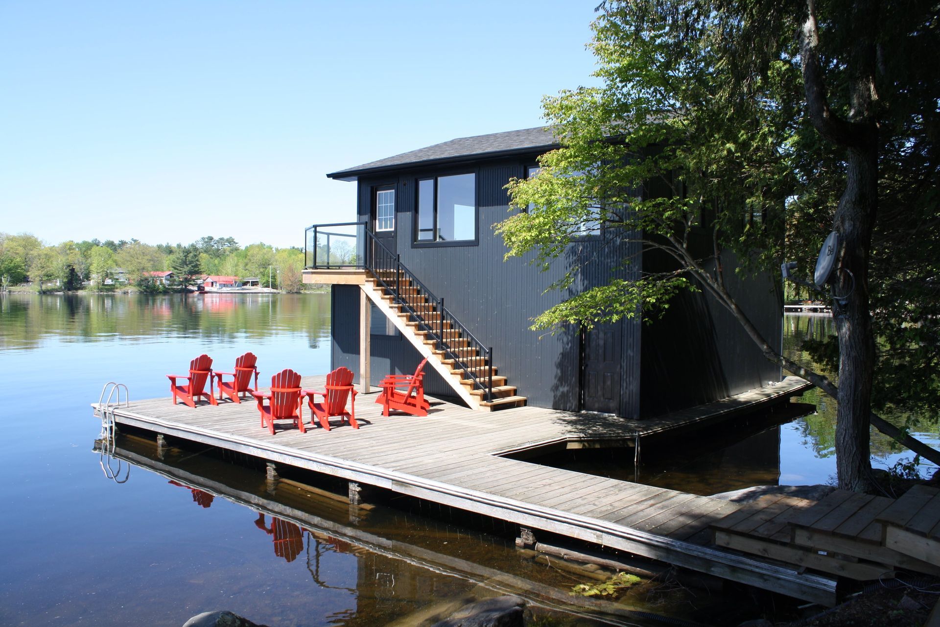 Black building with stairs and red Adirondack chairs on a dock over calm water.