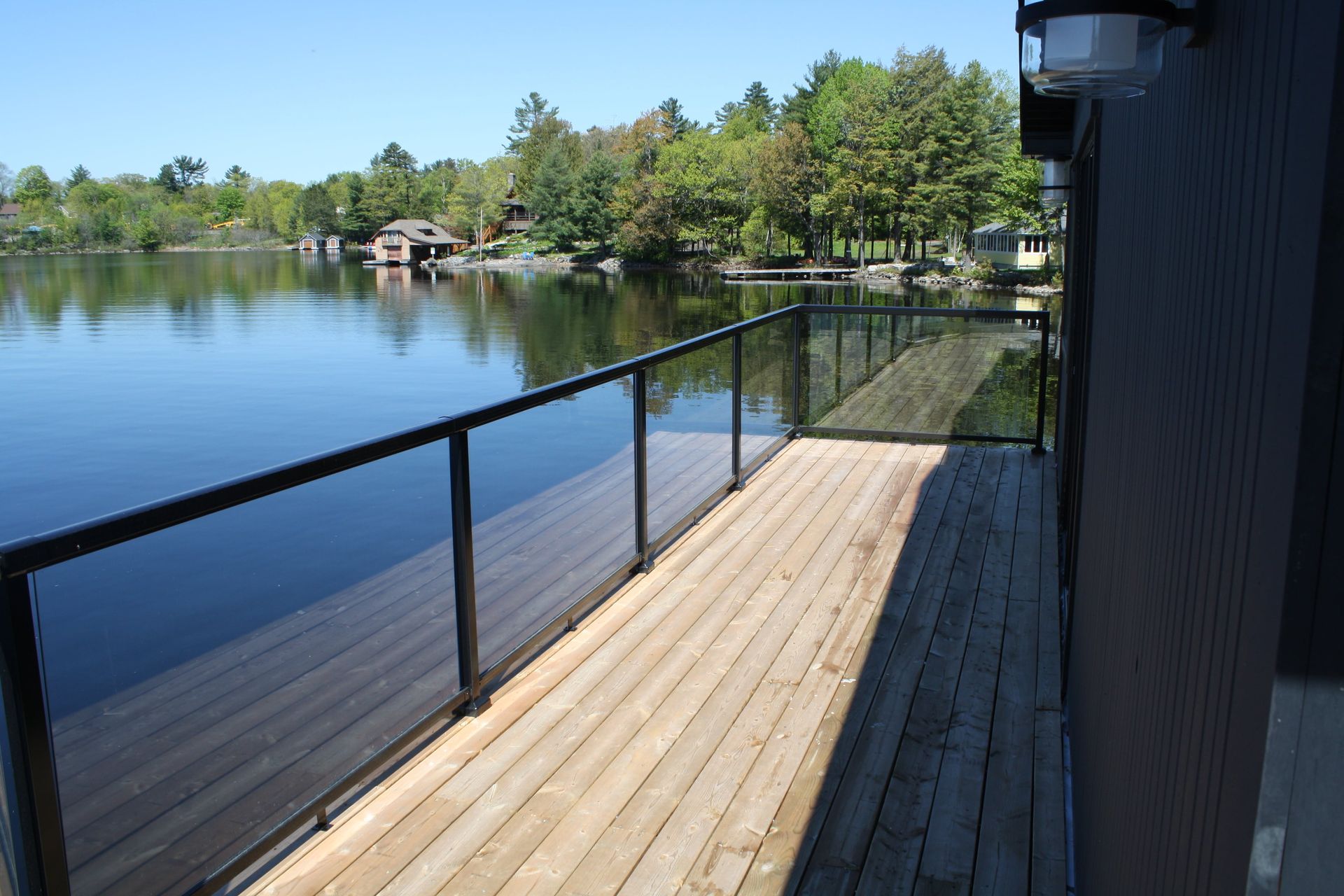 Wooden deck with glass railing overlooks calm lake, trees in the background.