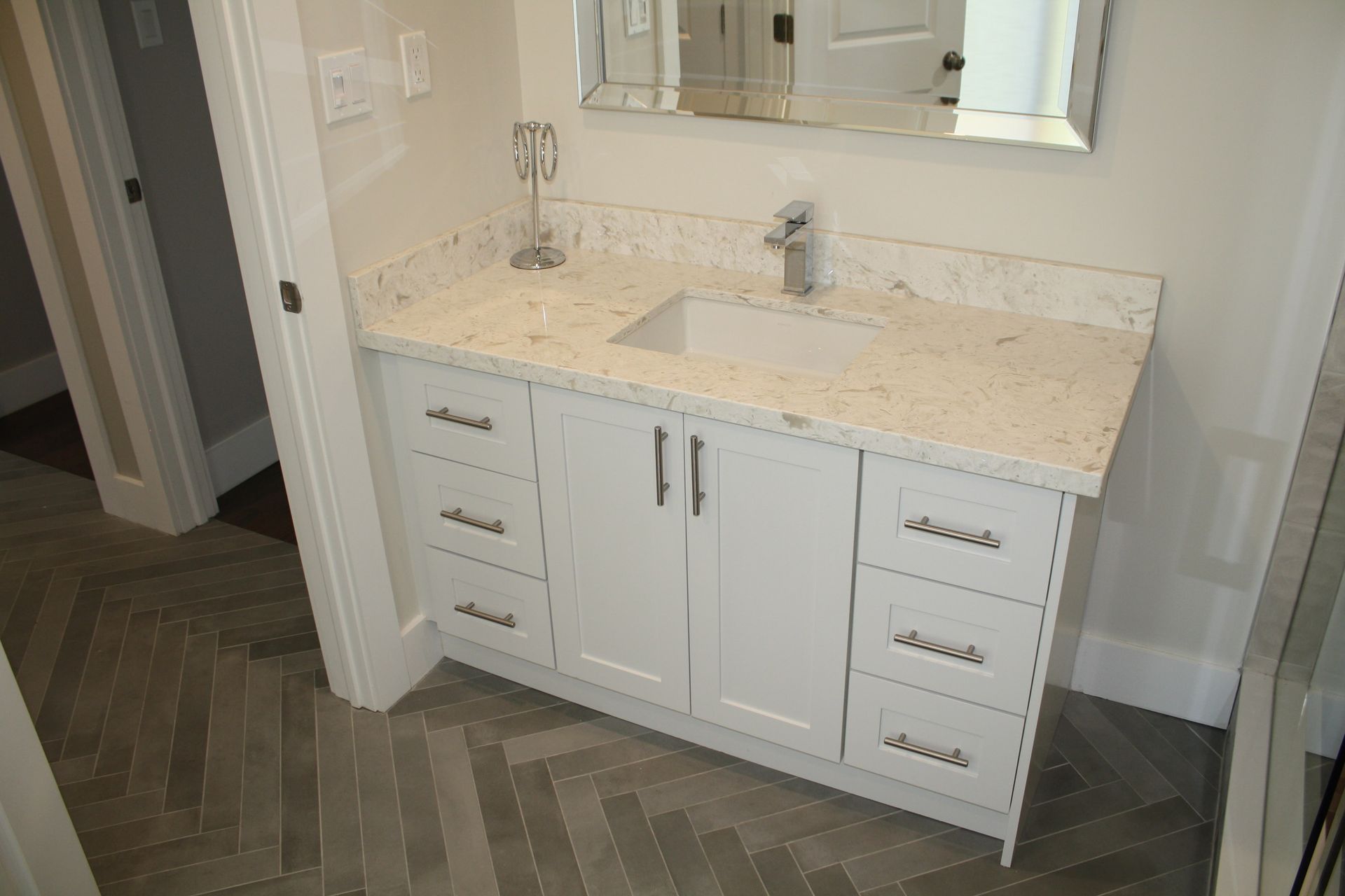 White bathroom vanity with marble countertop, sink, and mirror. Gray herringbone floor.