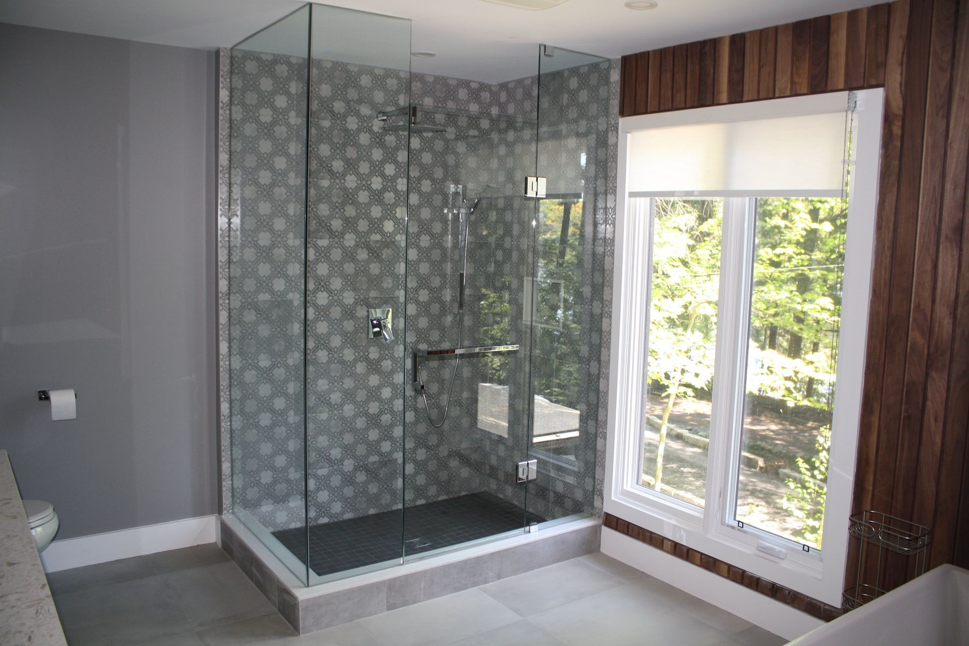 Glass shower in a modern bathroom with patterned tile and wood paneling near a window.