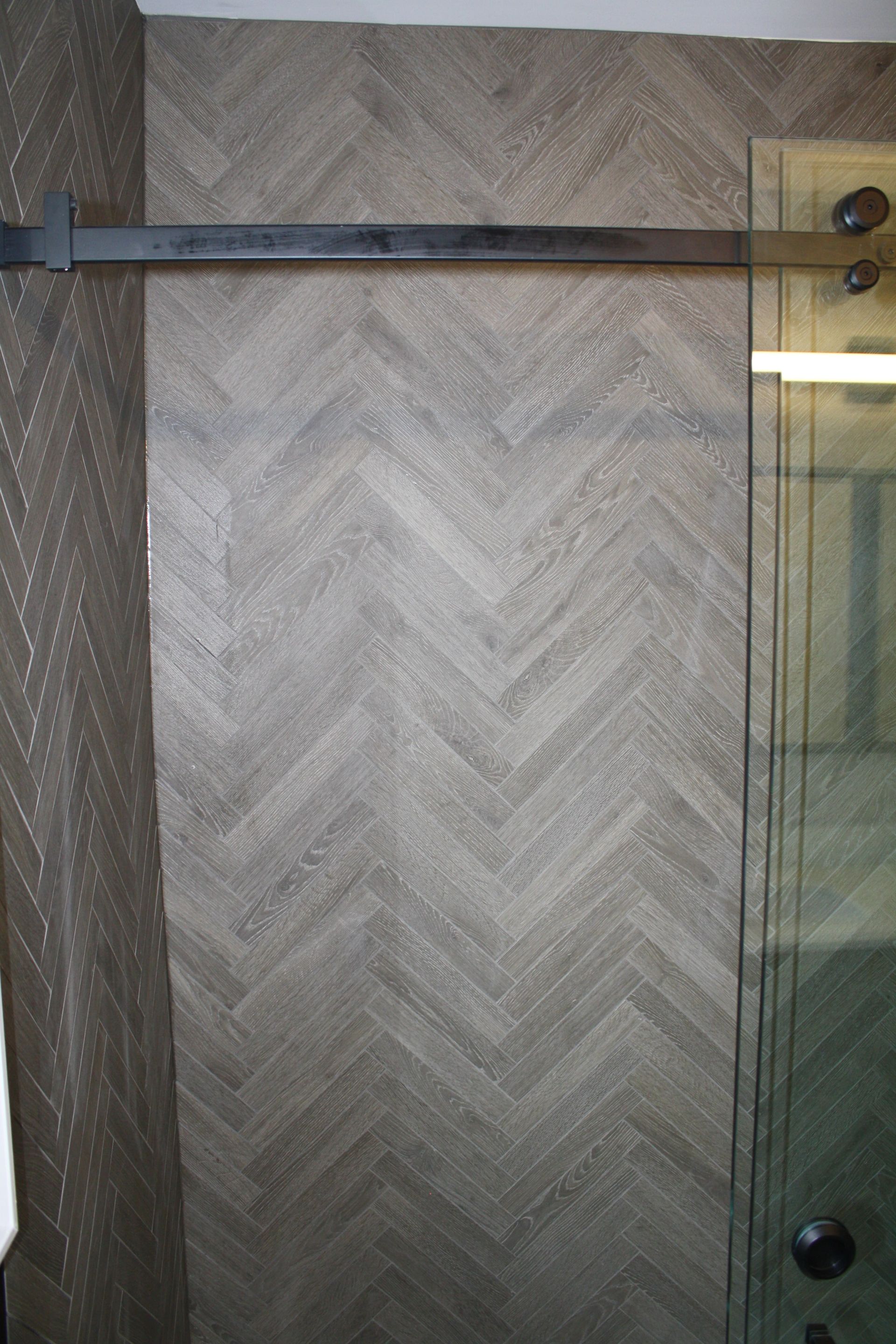 Gray herringbone-patterned wall in a shower, next to a glass door and black sliding hardware.