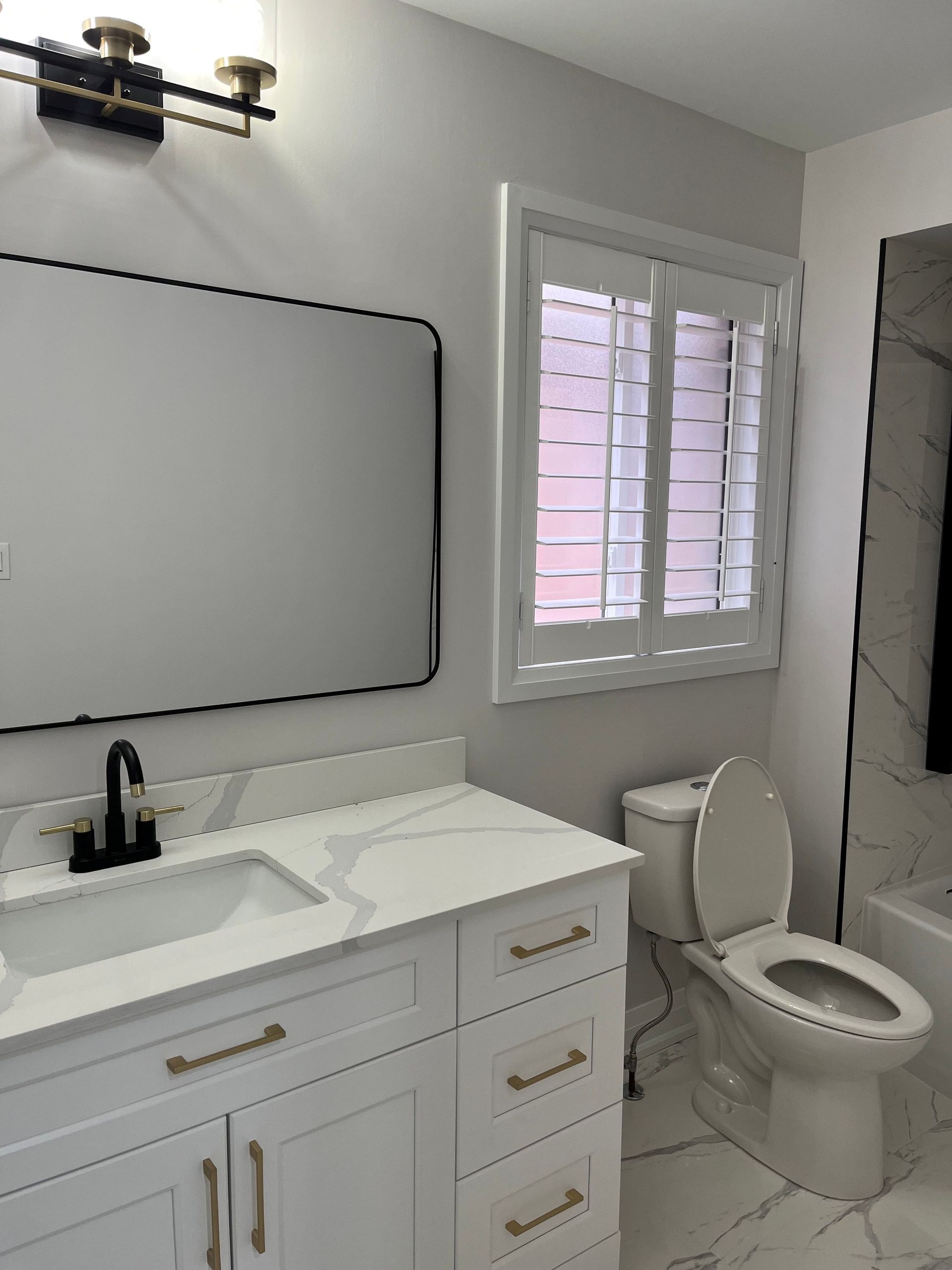 Bathroom with white vanity, marble countertop, black faucet, toilet, and window with shutters.