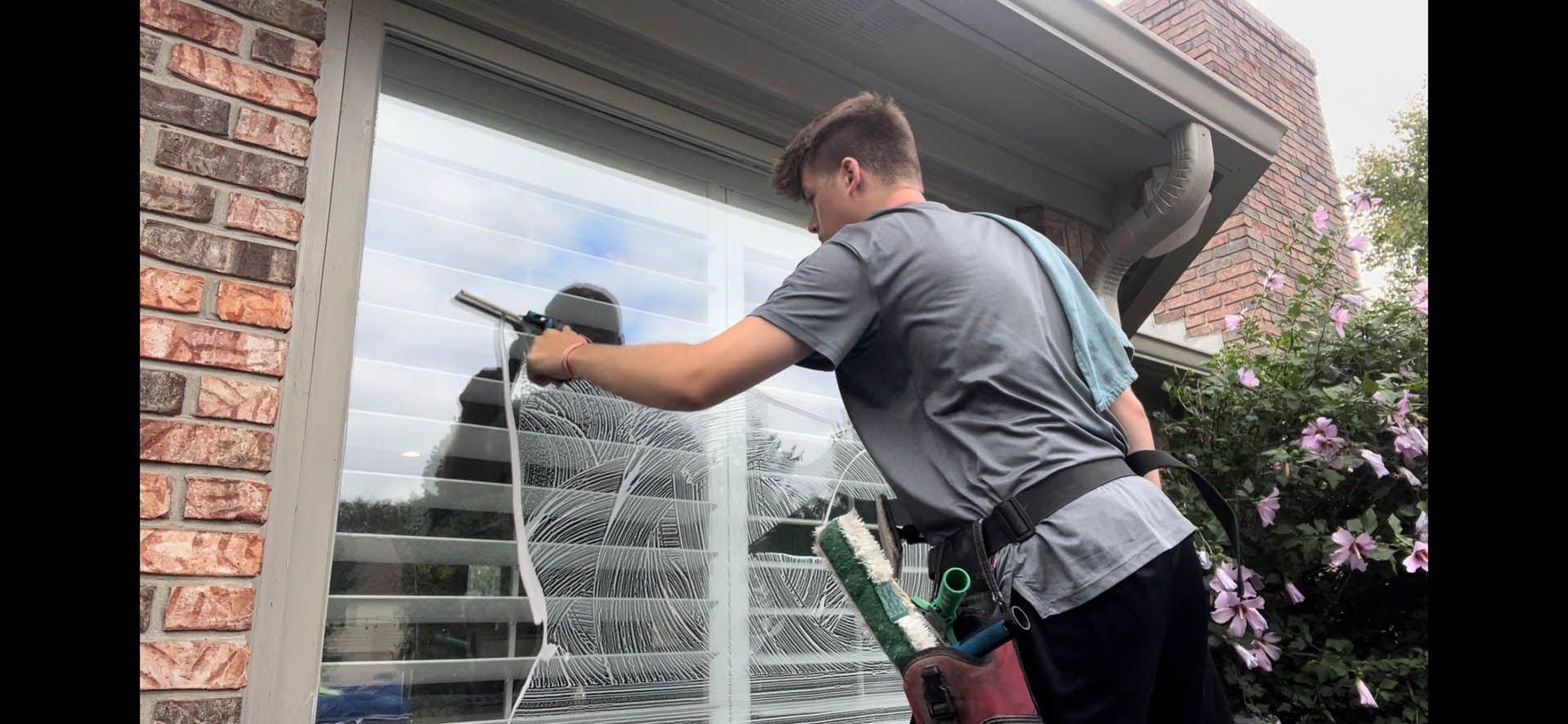 Worker pressure-washing a house exterior beside a brick wall and flowering bushes
