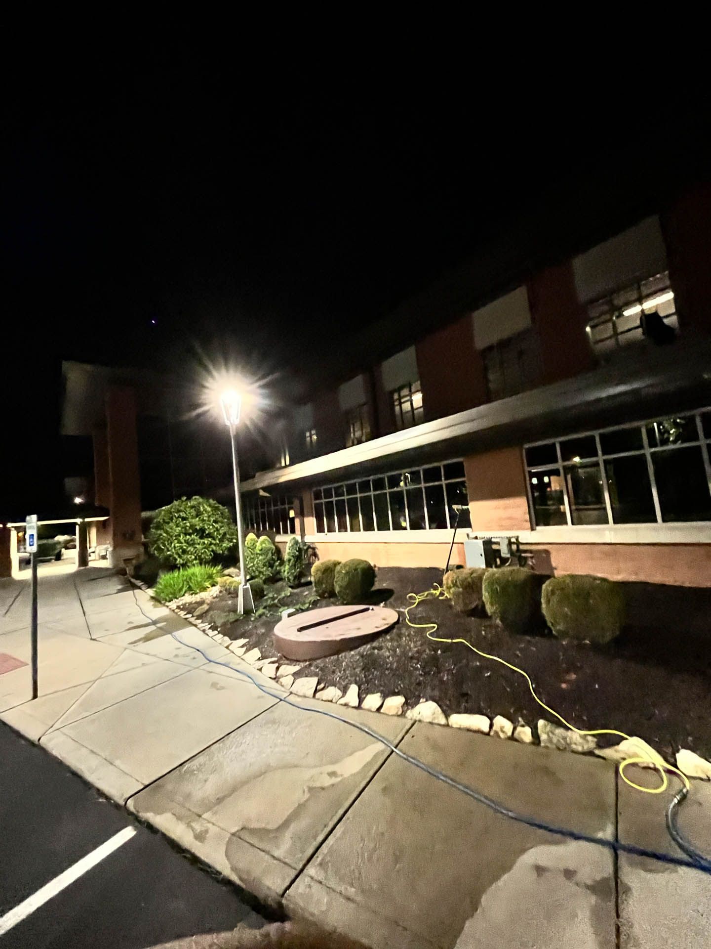 Nighttime exterior of a two-story building with lit windows, sidewalk, shrubs, and a bright streetlamp