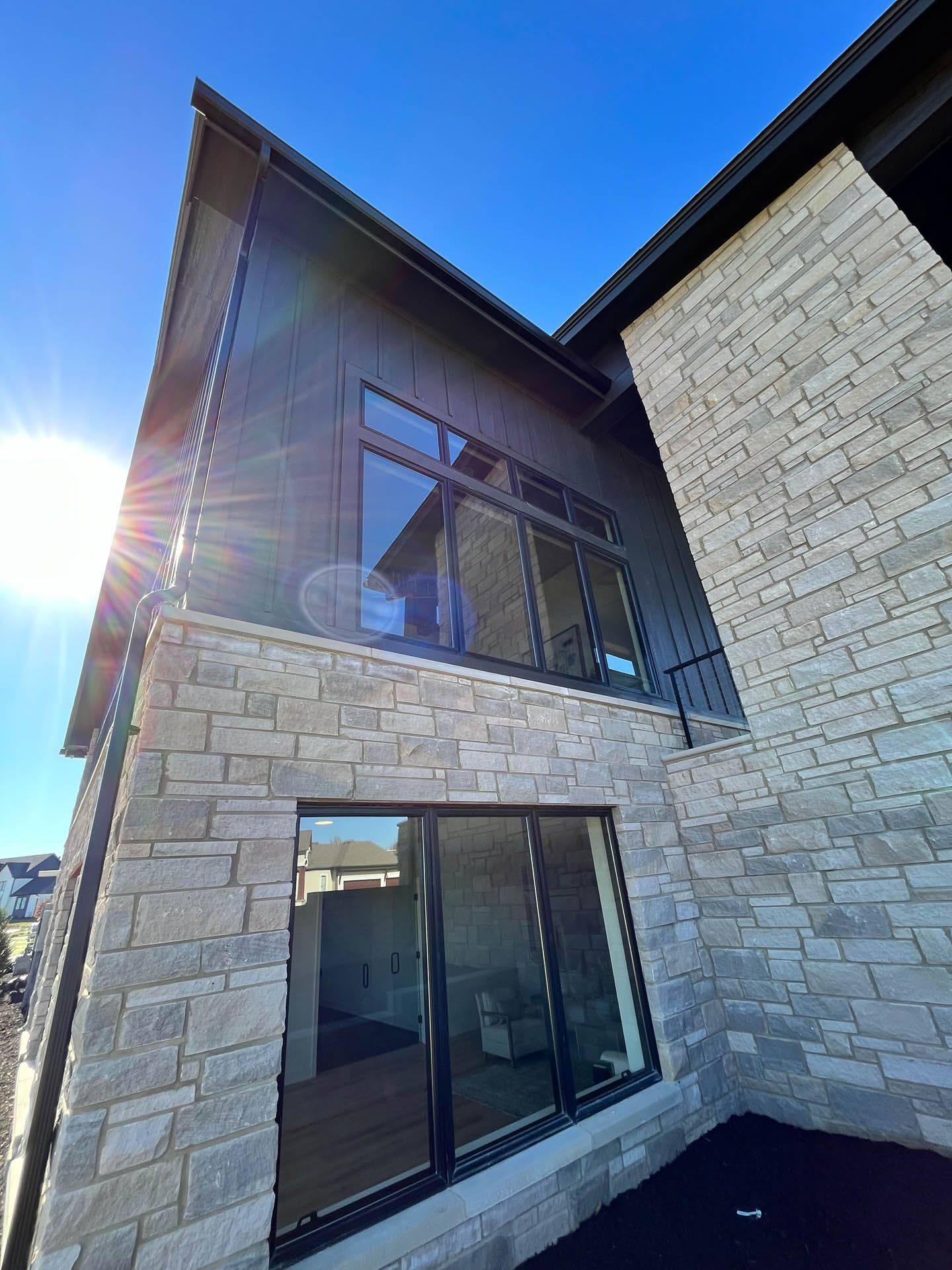 Modern stone house exterior with large windows and sun glare on a bright blue sky.
