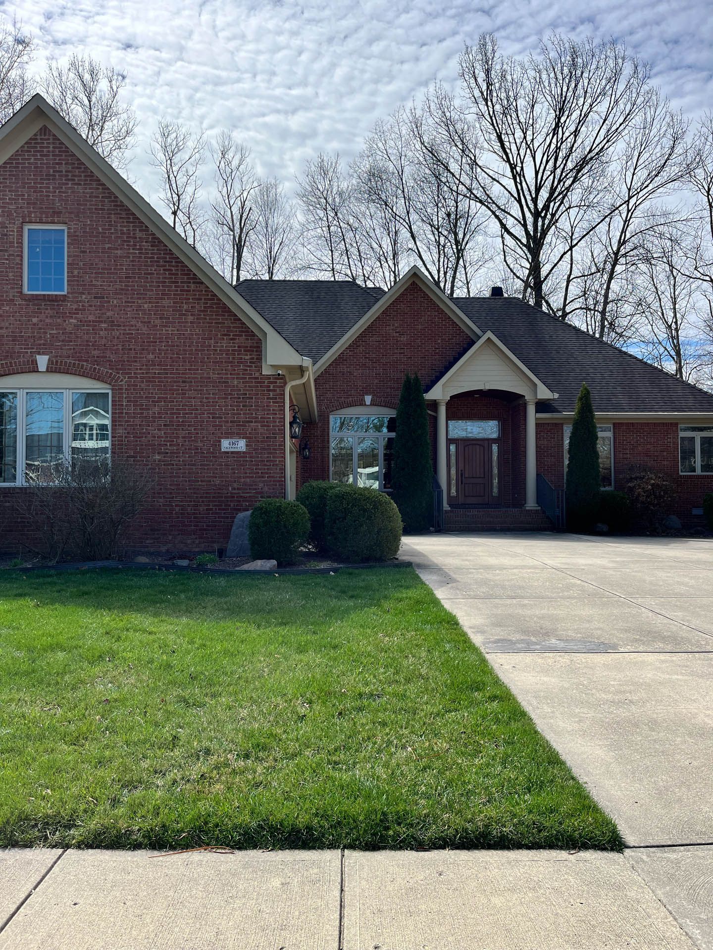 Brick suburban house with blue shutters, front lawn, and a concrete driveway under a partly cloudy sky