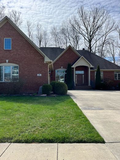 Brick suburban house with blue shutters, front lawn, and a concrete driveway under a partly cloudy sky