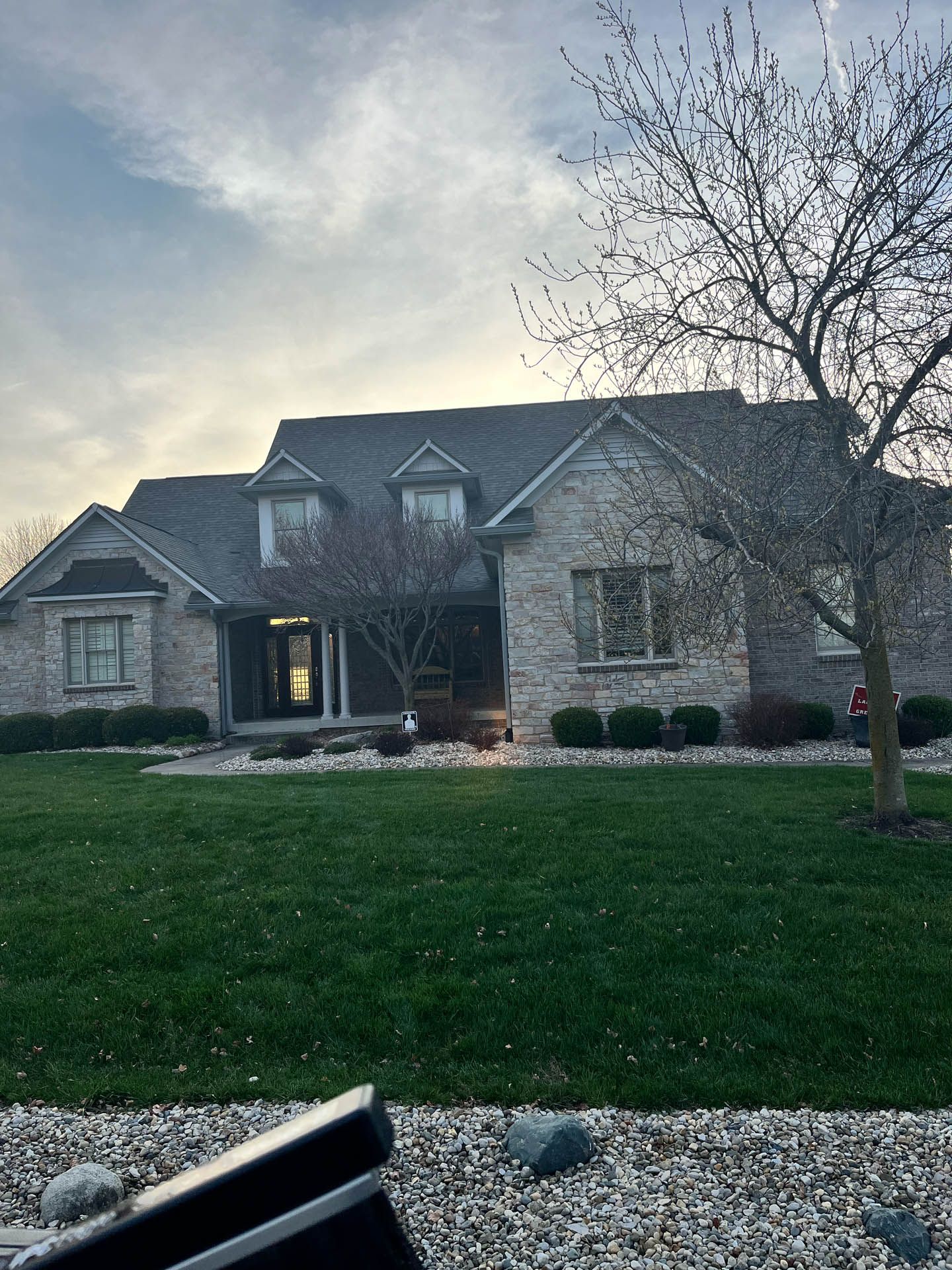 Stone house with lit porch at dusk, green lawn, bare tree, and cloudy sky.
