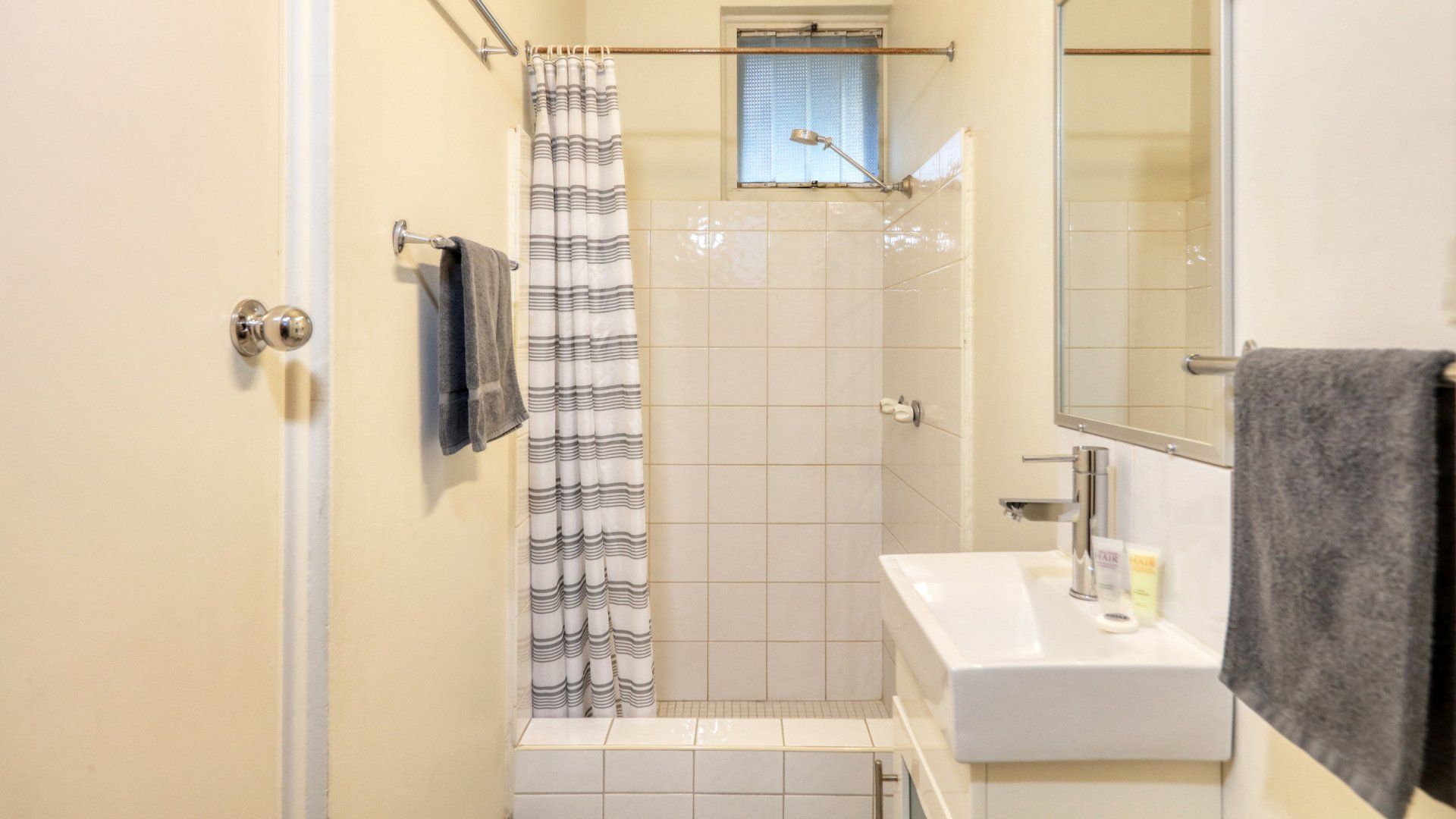 Small beige bathroom with a shower, white sink, mirror, and gray towels.