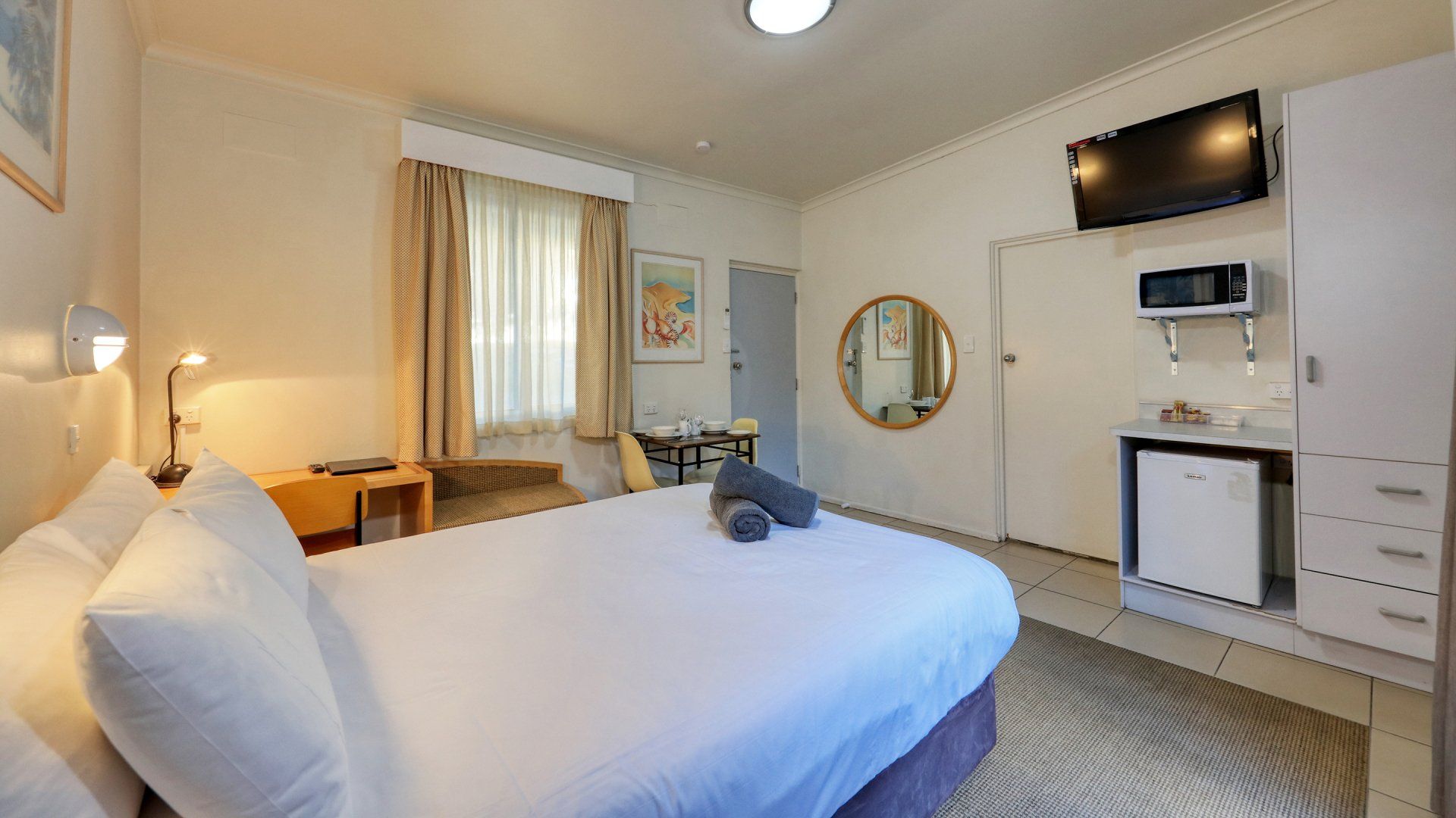 Bright hotel room with two beds, a window, wall-mounted TV, and a small desk and chair.