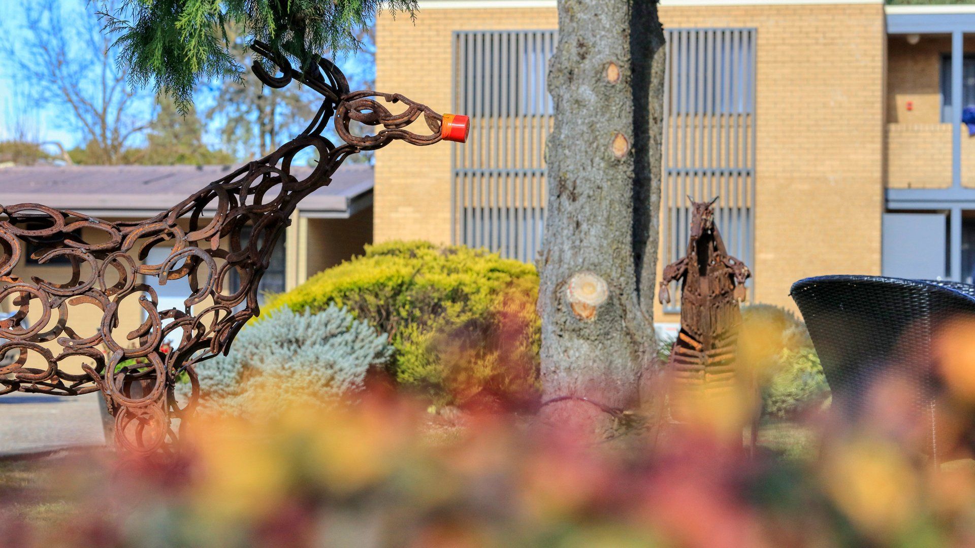 Abstract metal giraffe sculpture in a landscaped courtyard with bushes, a tree, and a building behind it