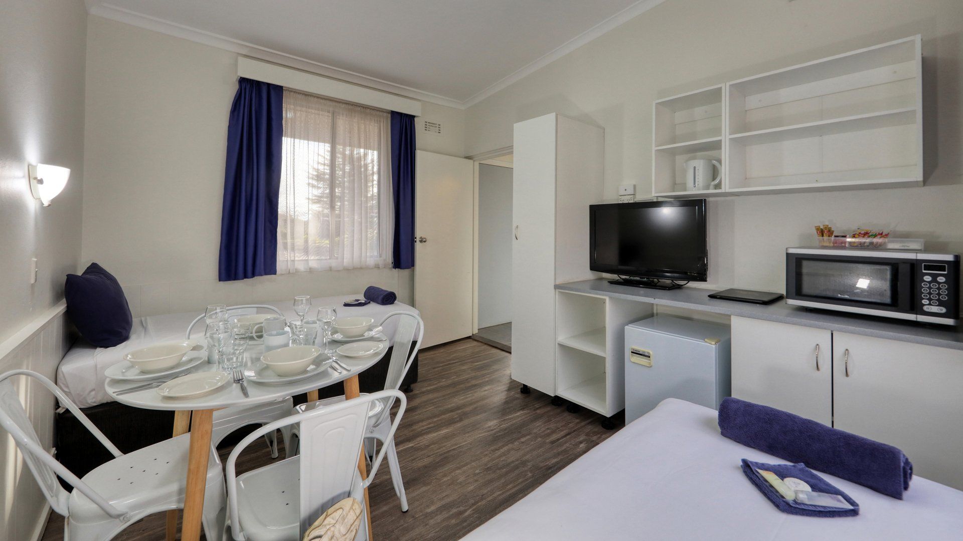 Bright studio apartment with bed, dining table, kitchenette, and TV, featuring white walls and blue accents