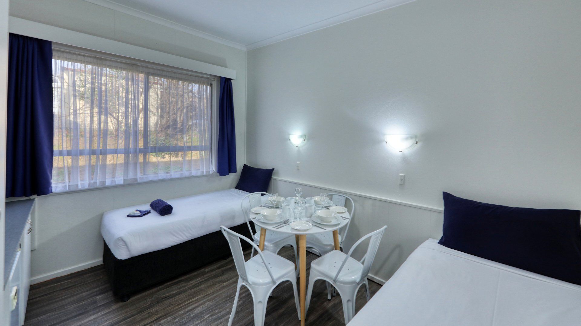 Bright twin bedroom with two single beds, a round table, and a large window with dark curtains.