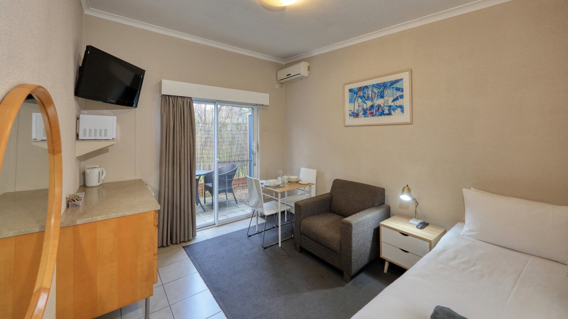 Small hotel room with bed, armchair, desk, TV, and balcony door with curtains
