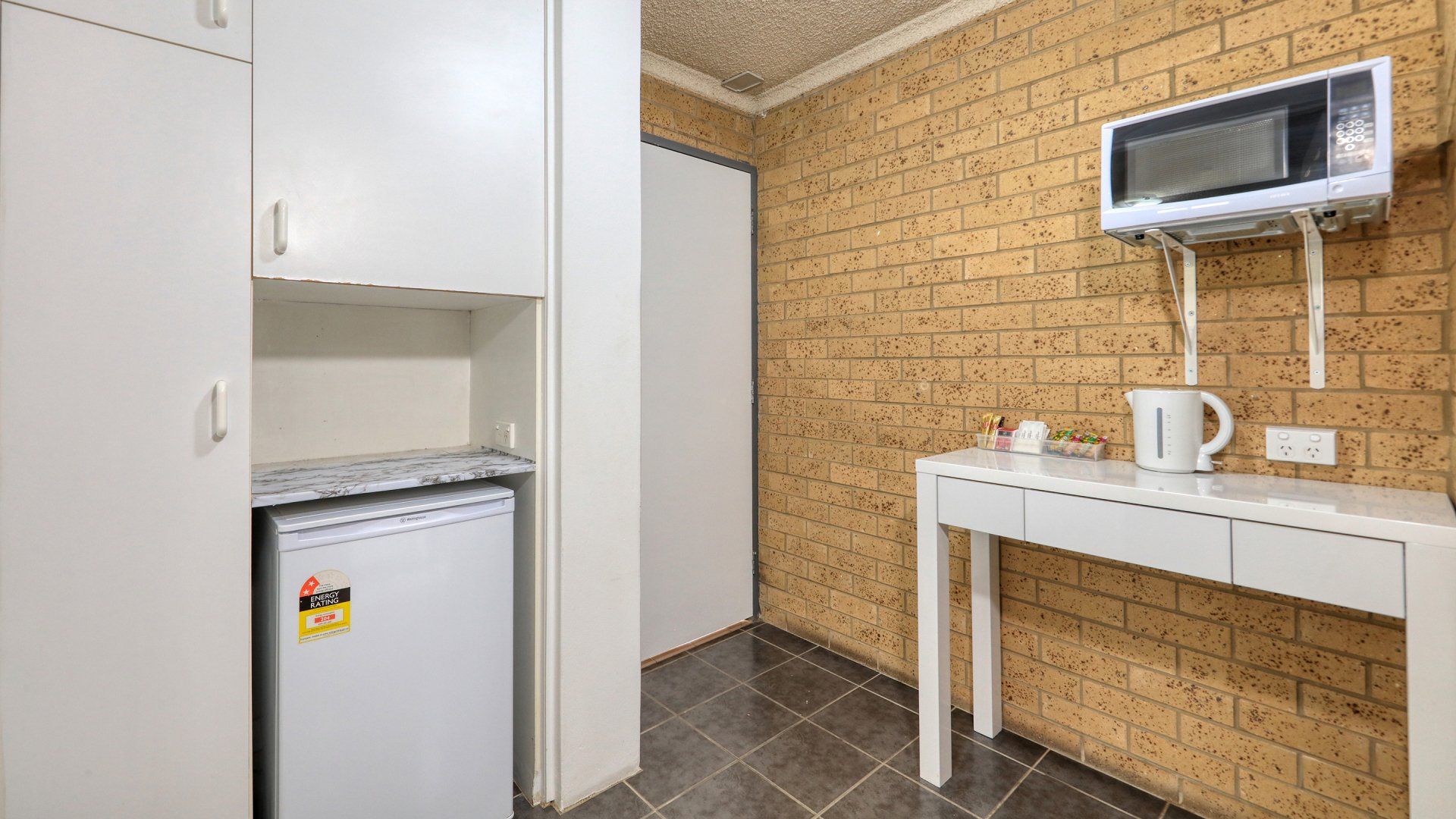 Small kitchenette with a white mini fridge, cabinet, table, and wall-mounted microwave on a brick wall.