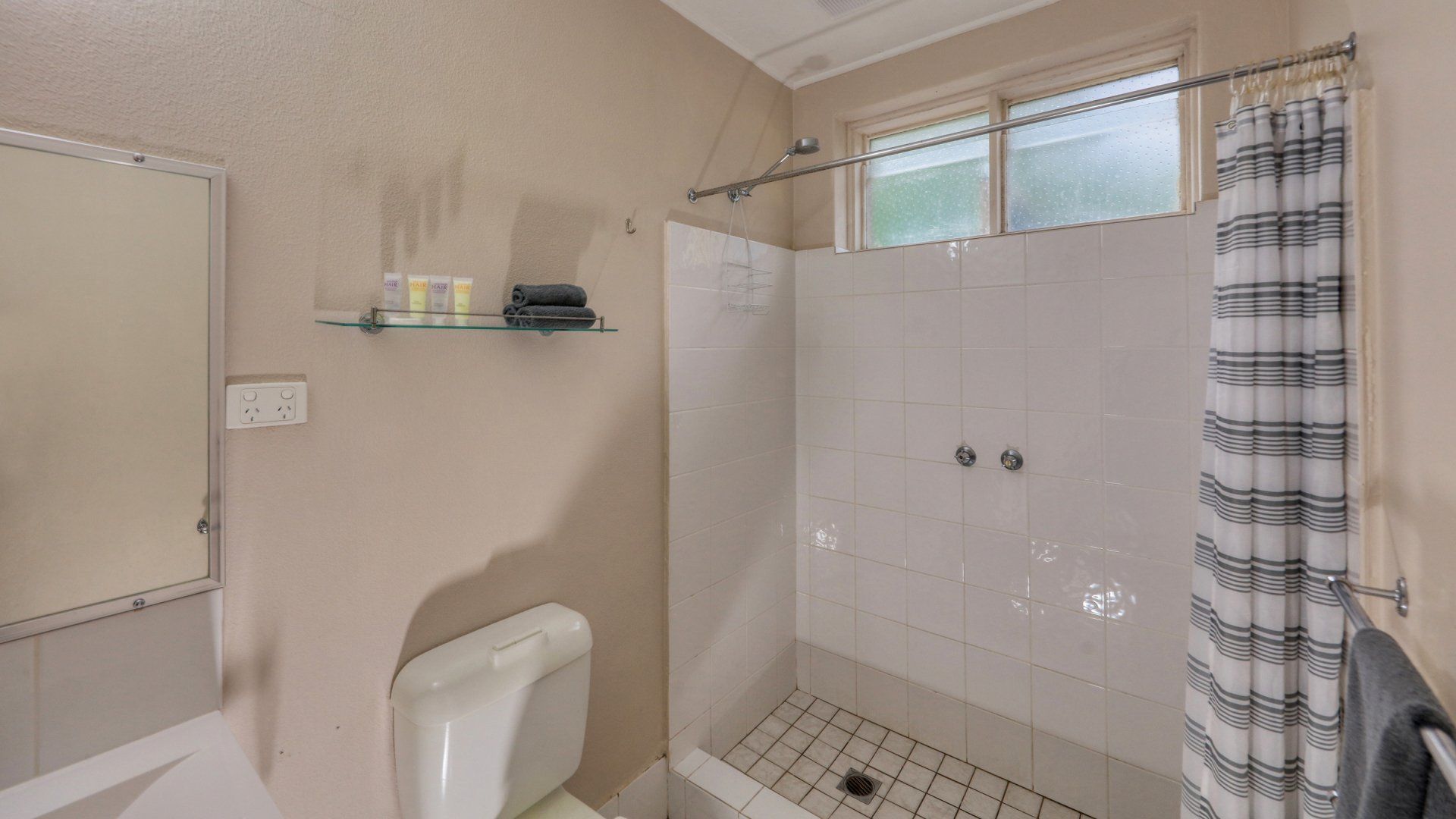Small beige bathroom with a toilet, wall shelf, shower stall, and striped curtain.